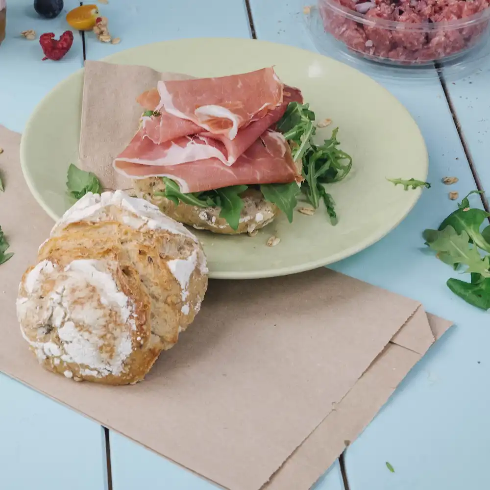 Scheiben Prosciutto auf einem grünen Teller mit Rucola auf einem braunen Brötchen; ein runder, luftiger Brötchen liegt daneben.