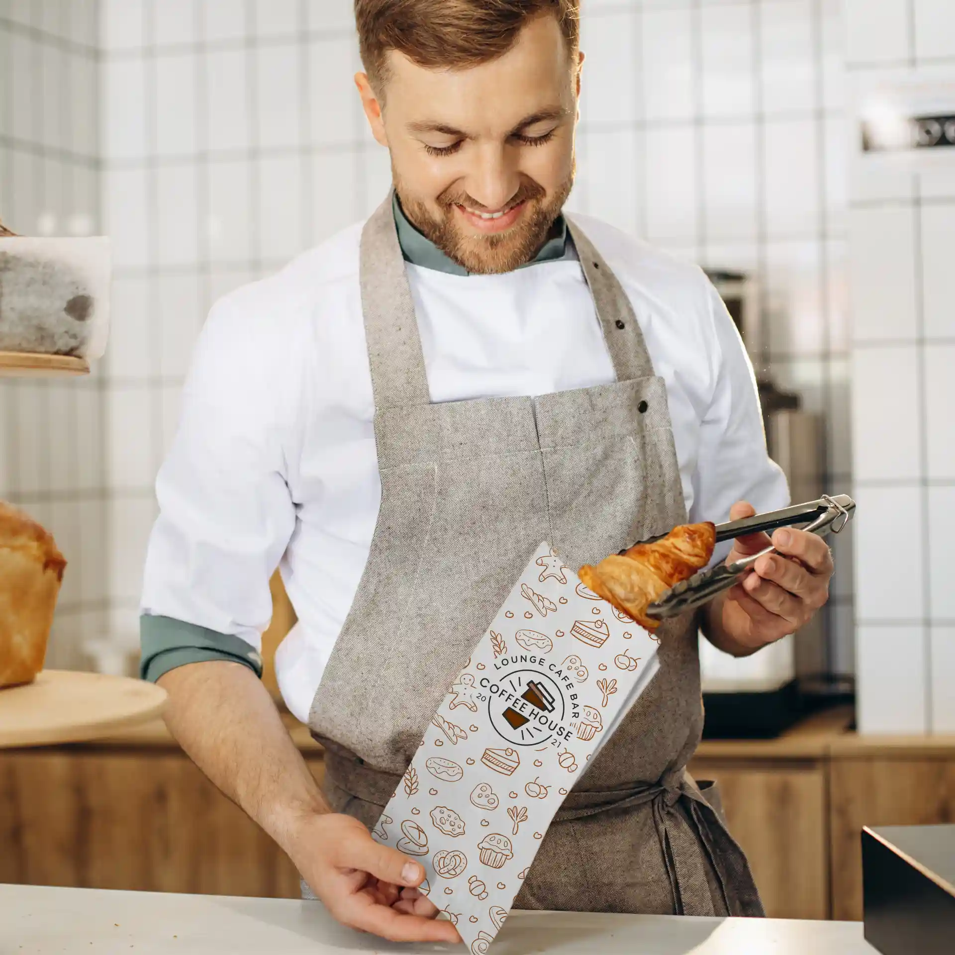 Bild eines Mannes in einer Schürze, der eine bedruckte Papiertüte mit einem Croissant in einem Café hält.