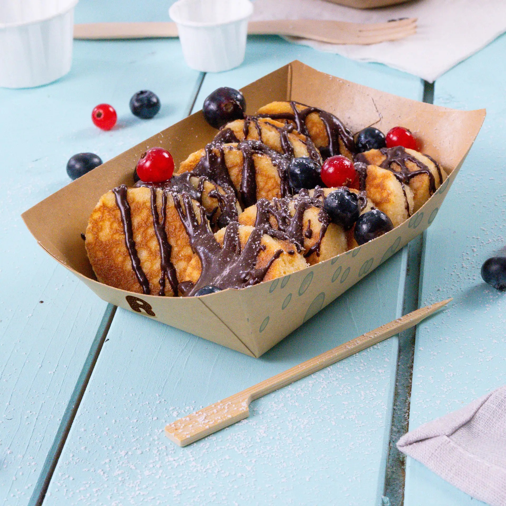 A brown cardboard snack tray filled with chocolate-drizzled desserts, topped with red cherries and black blueberries, on a blue surface.