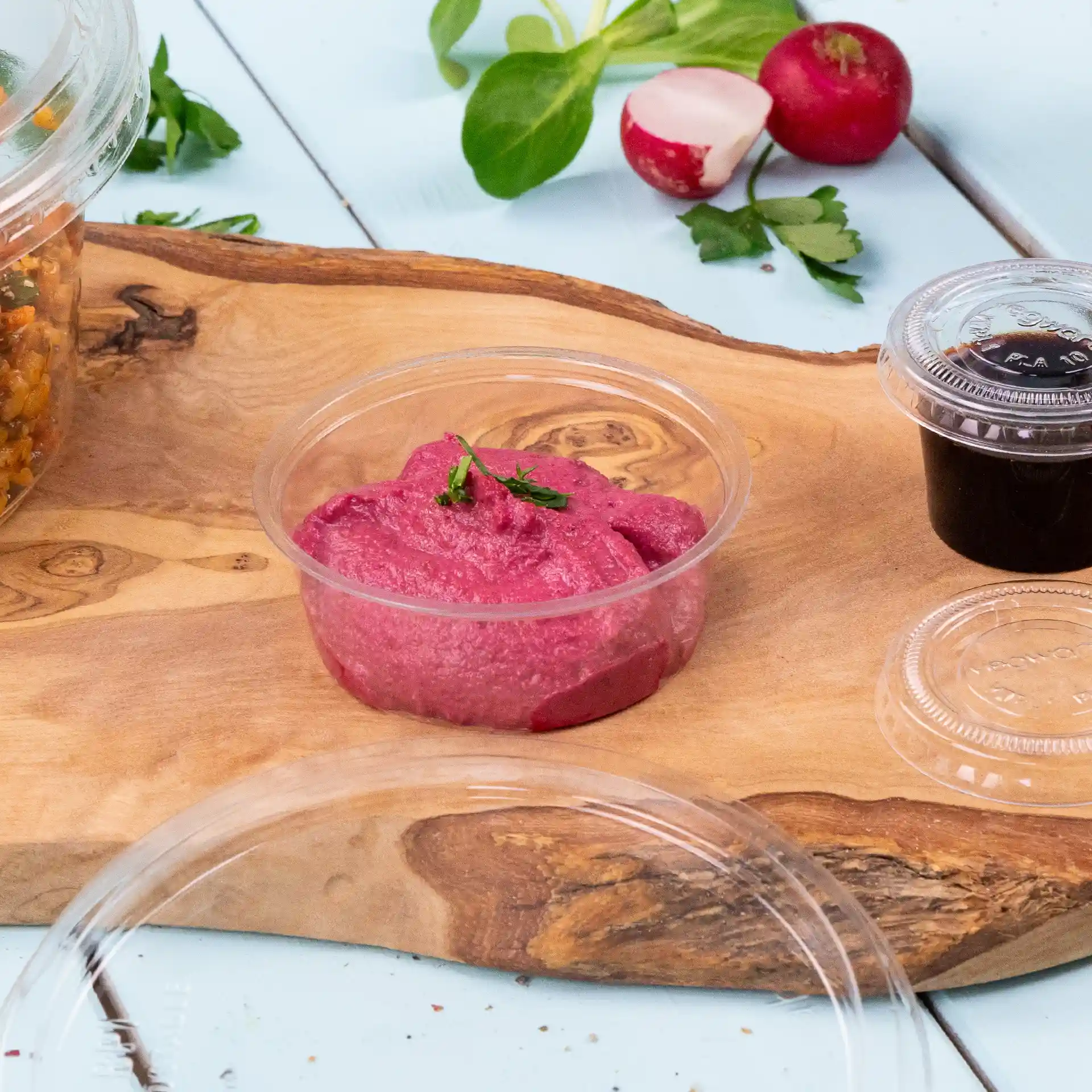 Clear round dip pot with pink dip, garnished with a small green herb, on a wooden surface with vegetables.