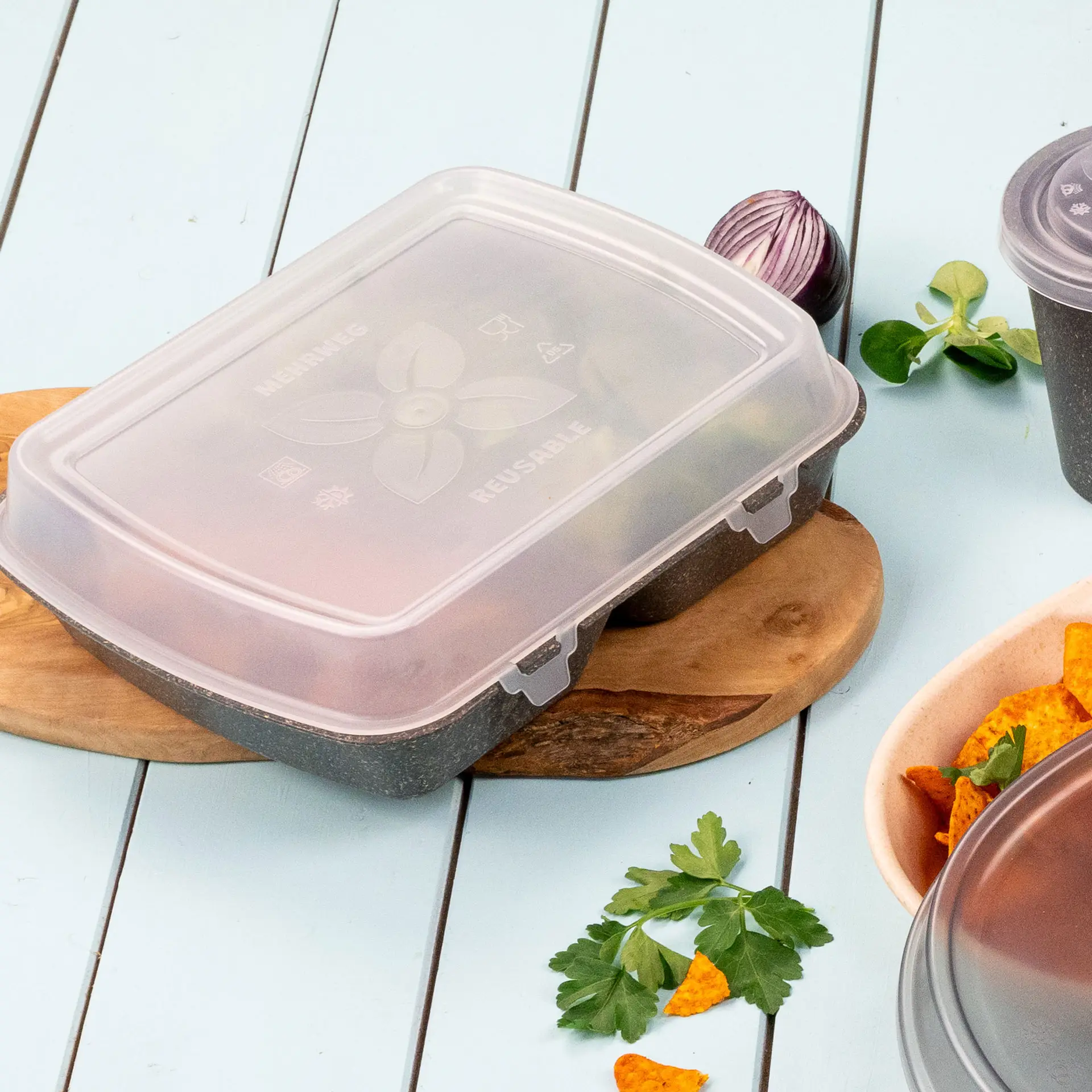 Transparente wiederverwendbare Lebensmittelbox mit Deckel, auf einem Holzbrett, umgeben von Kräutern und Snacks.
