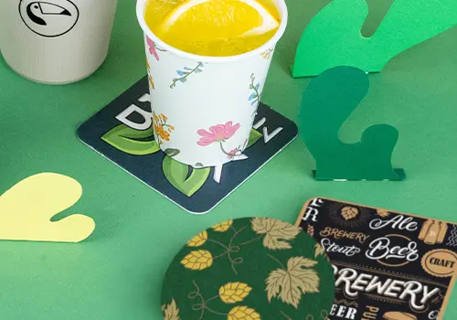 Stacks of drink coasters on a wooden surface; some feature a logo with a bird and others have a design with beer and text.