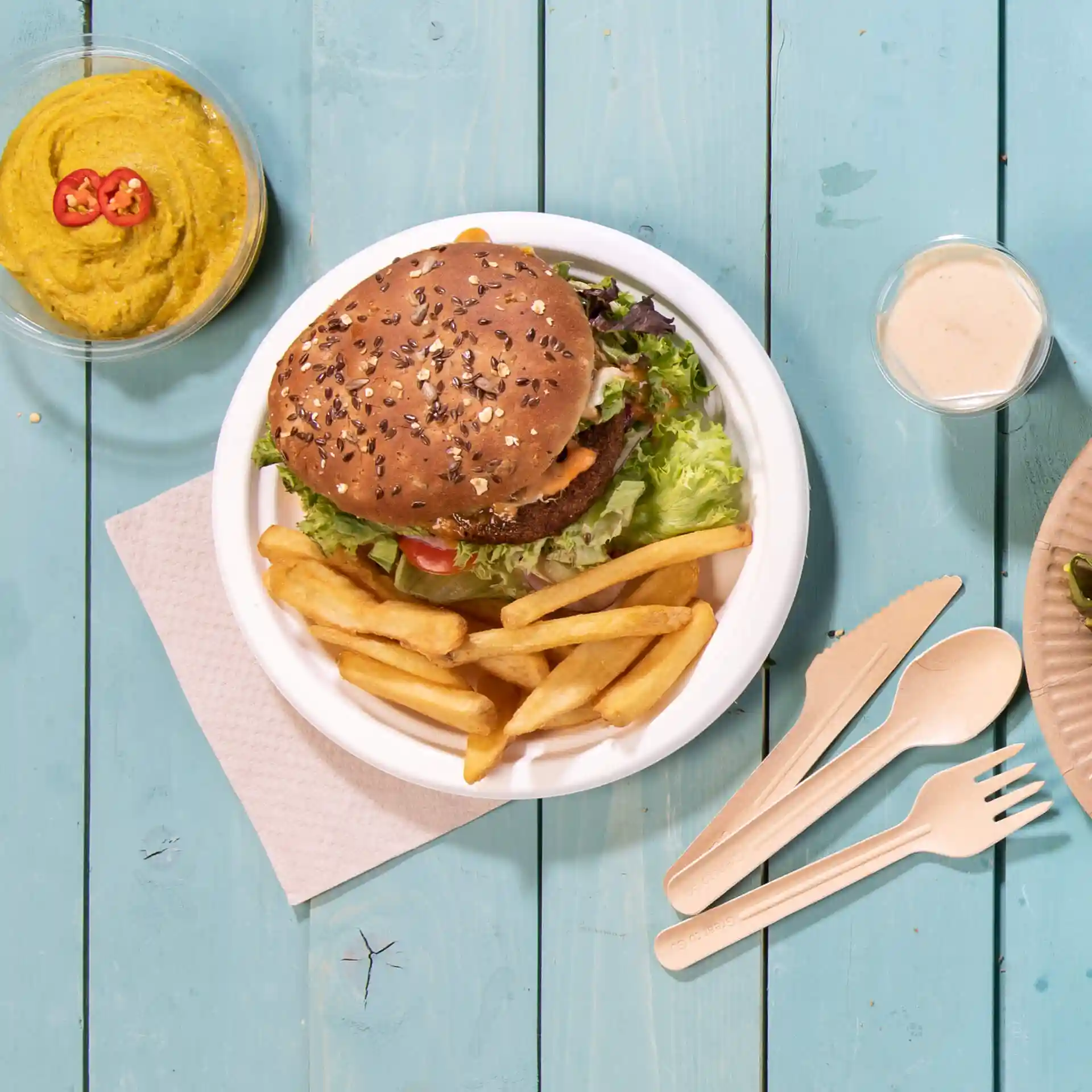 Bild eines runden weißen Zuckerröhren-Tellers mit einem Burger, Pommes und einer Schüssel Hummus auf einer blauen Holzoberfläche.