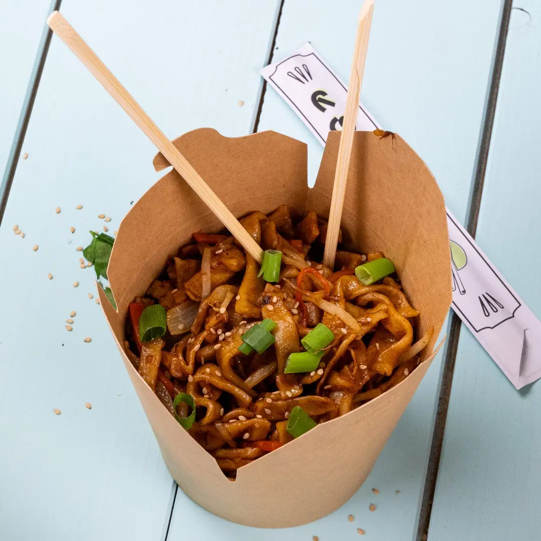Brown paper container filled with stir-fried noodles, topped with green onions and sesame seeds, with chopsticks inserted.
