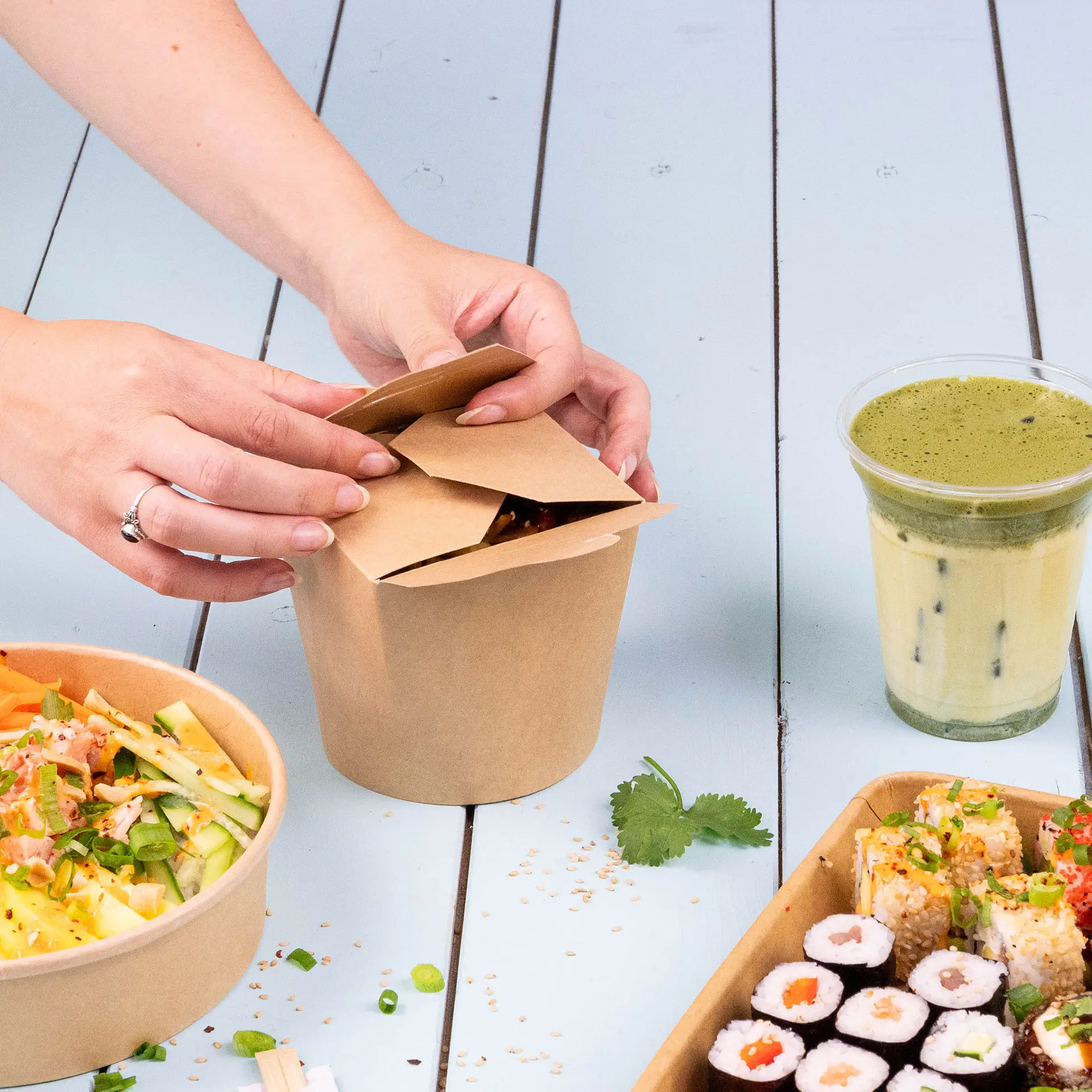A brown noodle box filled with fried noodles, topped with sliced red chilies and cilantro, on a bamboo mat with other food containers.