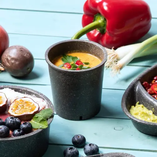 Wiederverwendbare Suppenschale gefüllt mit Suppe und mit Kräutern garniert, umgeben von frischem Gemüse und Obst auf einer blauen Fläche.