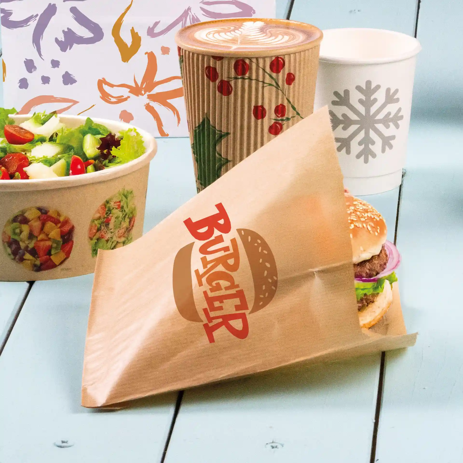 Braune bedruckte Burgerhülle mit sichtbarem Burger, Schüssel mit Salat und Kaffeebecher im Hintergrund.