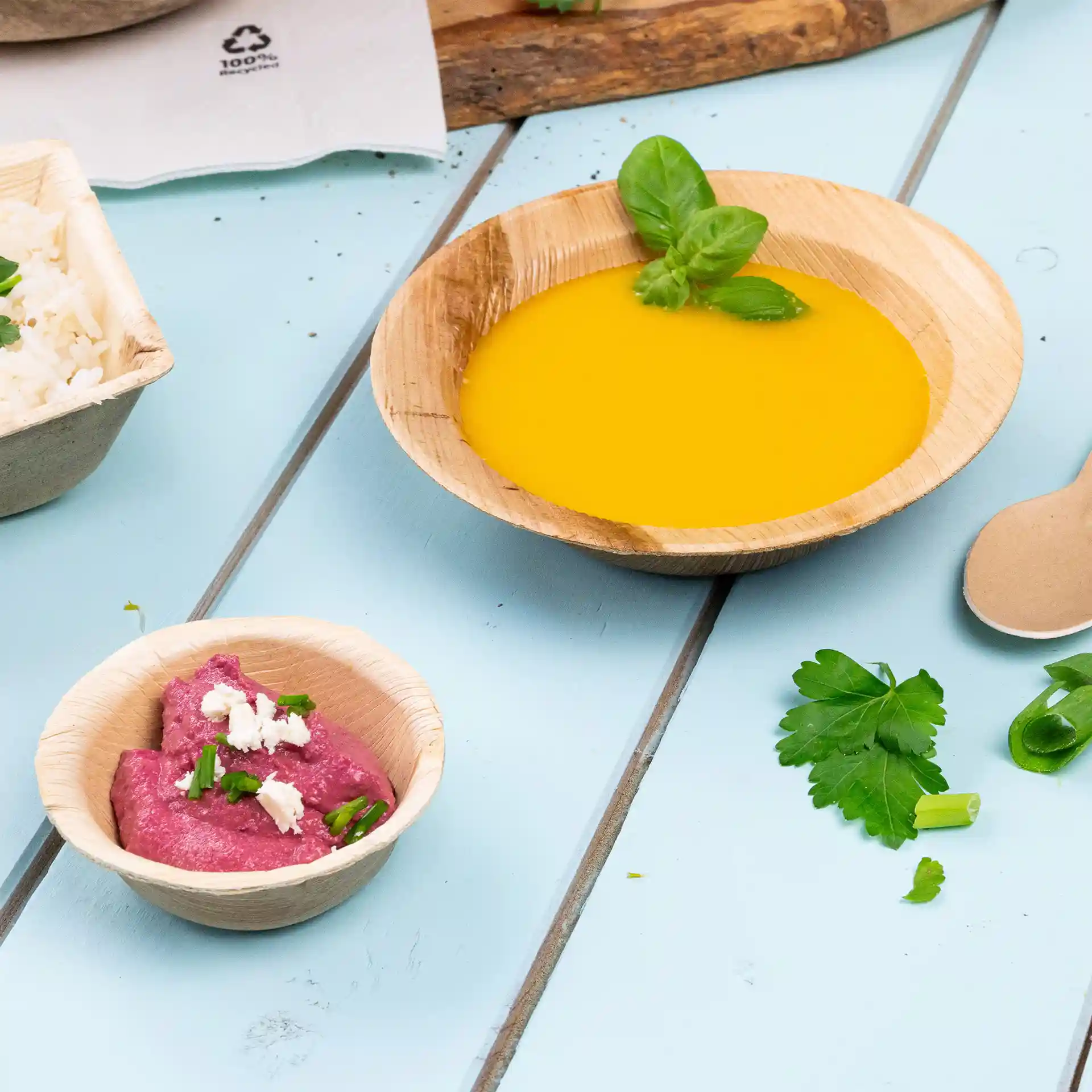Bowl made of palm leaf containing yellow liquid and garnished with a basil leaf, alongside a smaller bowl of pink spread.