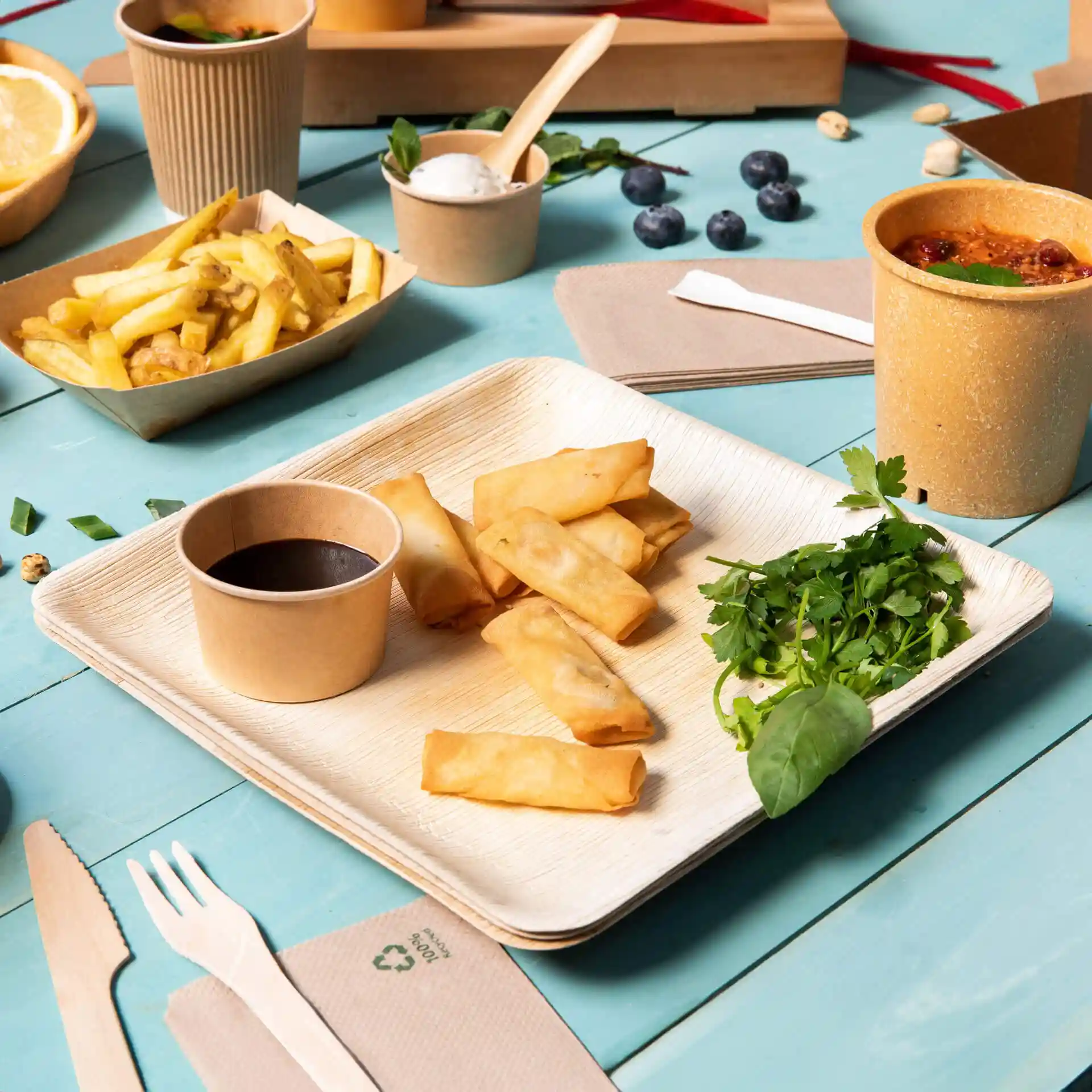 Square palm leaf plate with spring rolls, a cup of sauce, fresh herbs, and side dishes on a blue table.