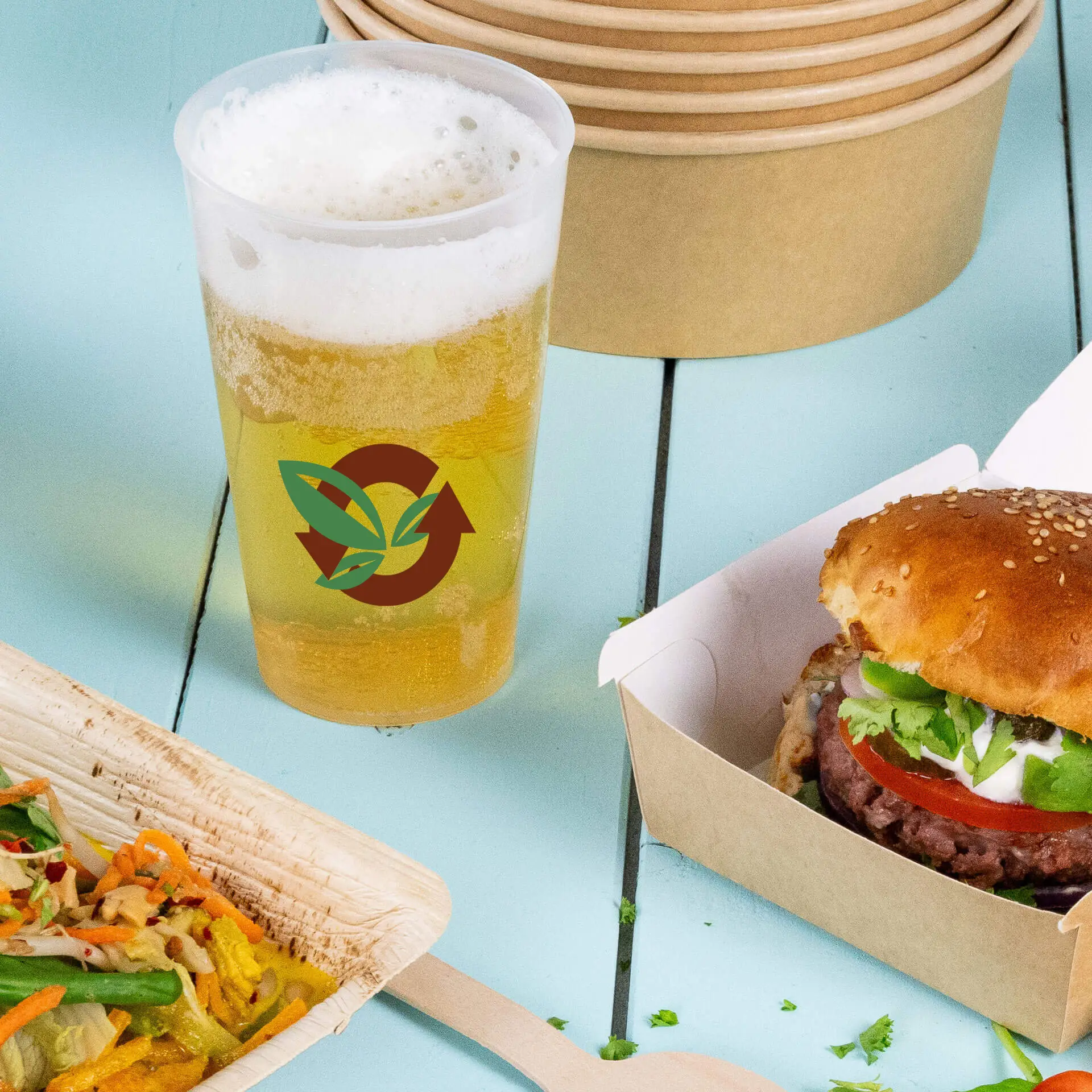 Reusable cup with logo containing beer, alongside a sandwich and a wooden fork on a blue surface.