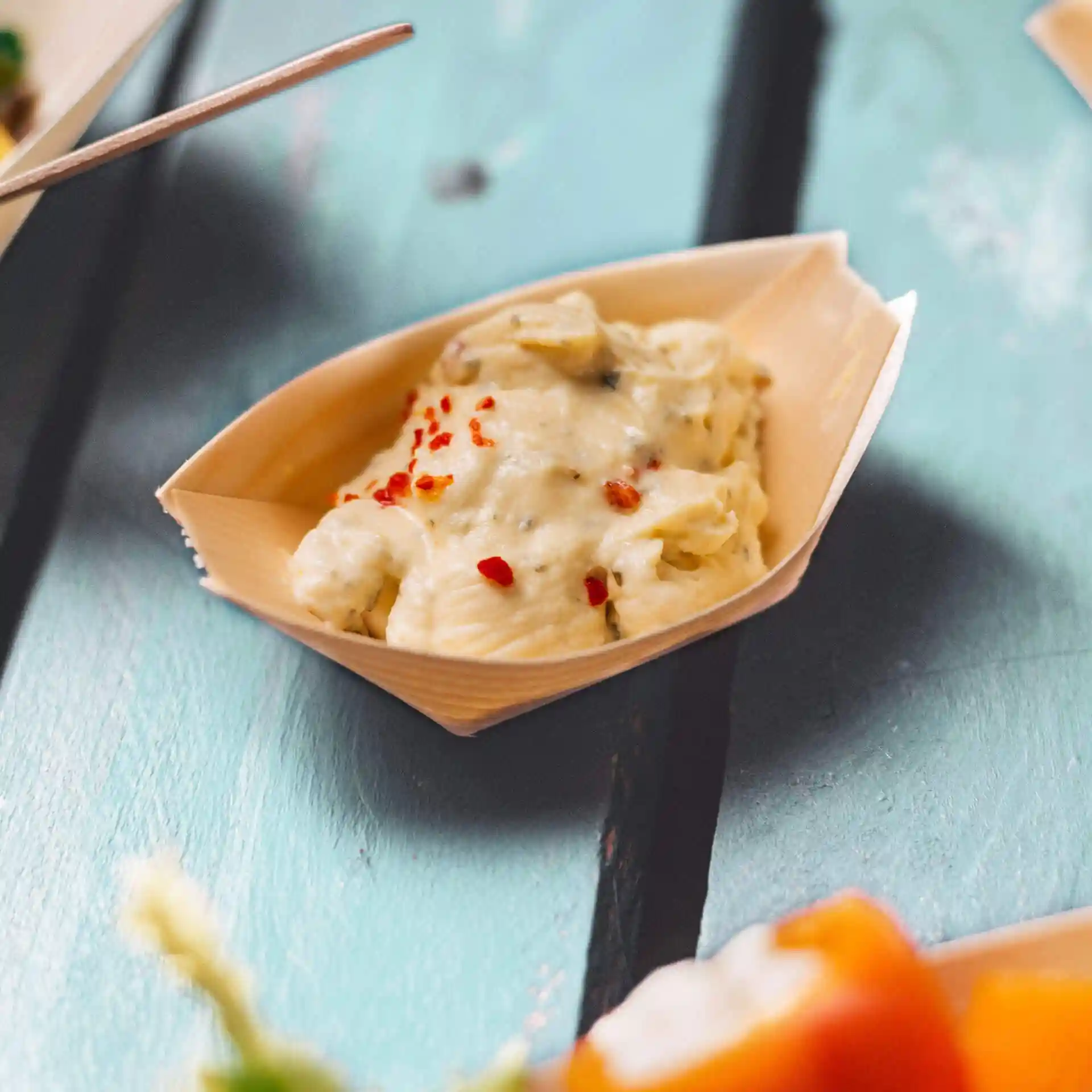Wooden boat-shaped dish containing creamy food topped with red pepper flakes on a blue wooden surface.