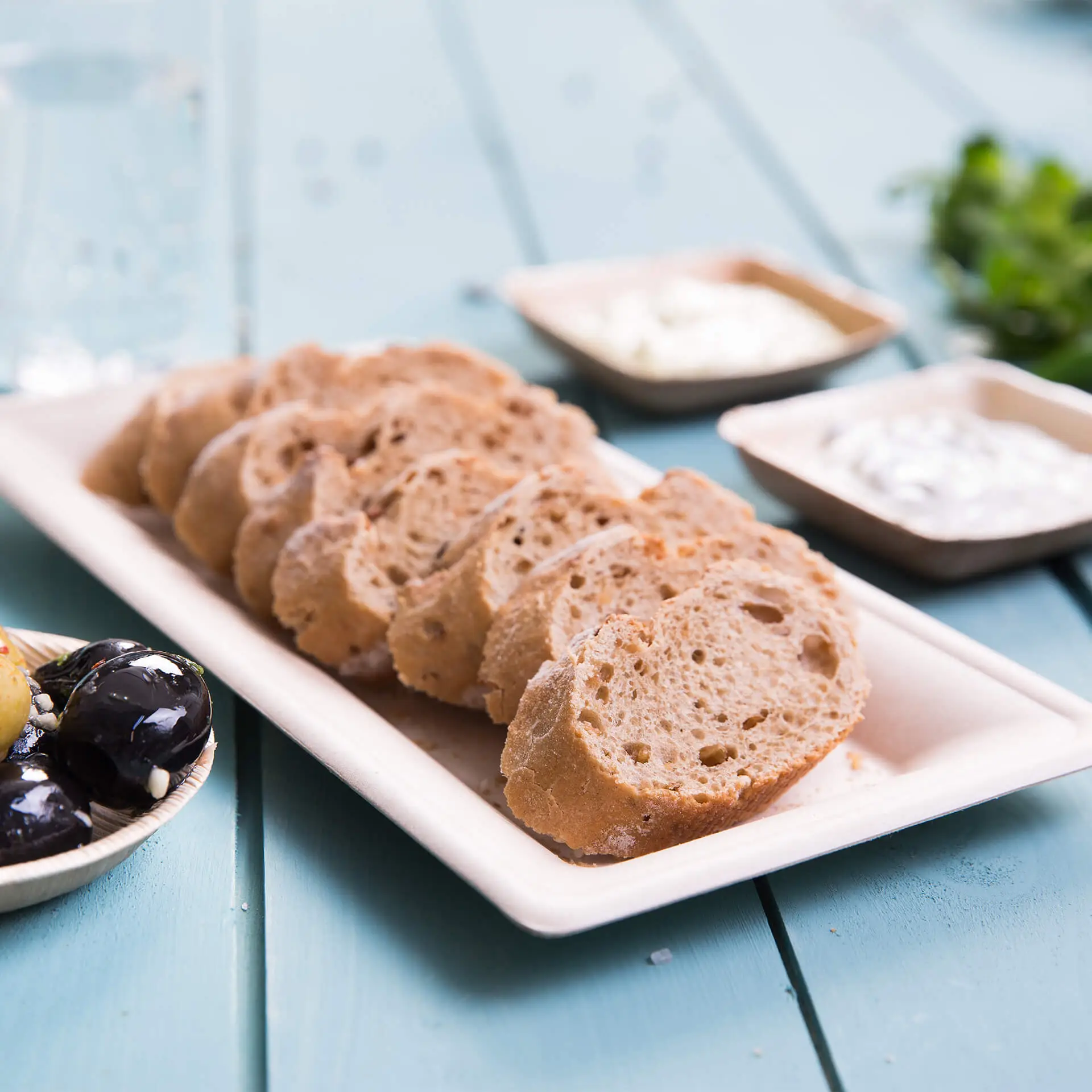 Sliced bread on a rectangular biodegradable sugarcane plate with condiments and olives in the background.