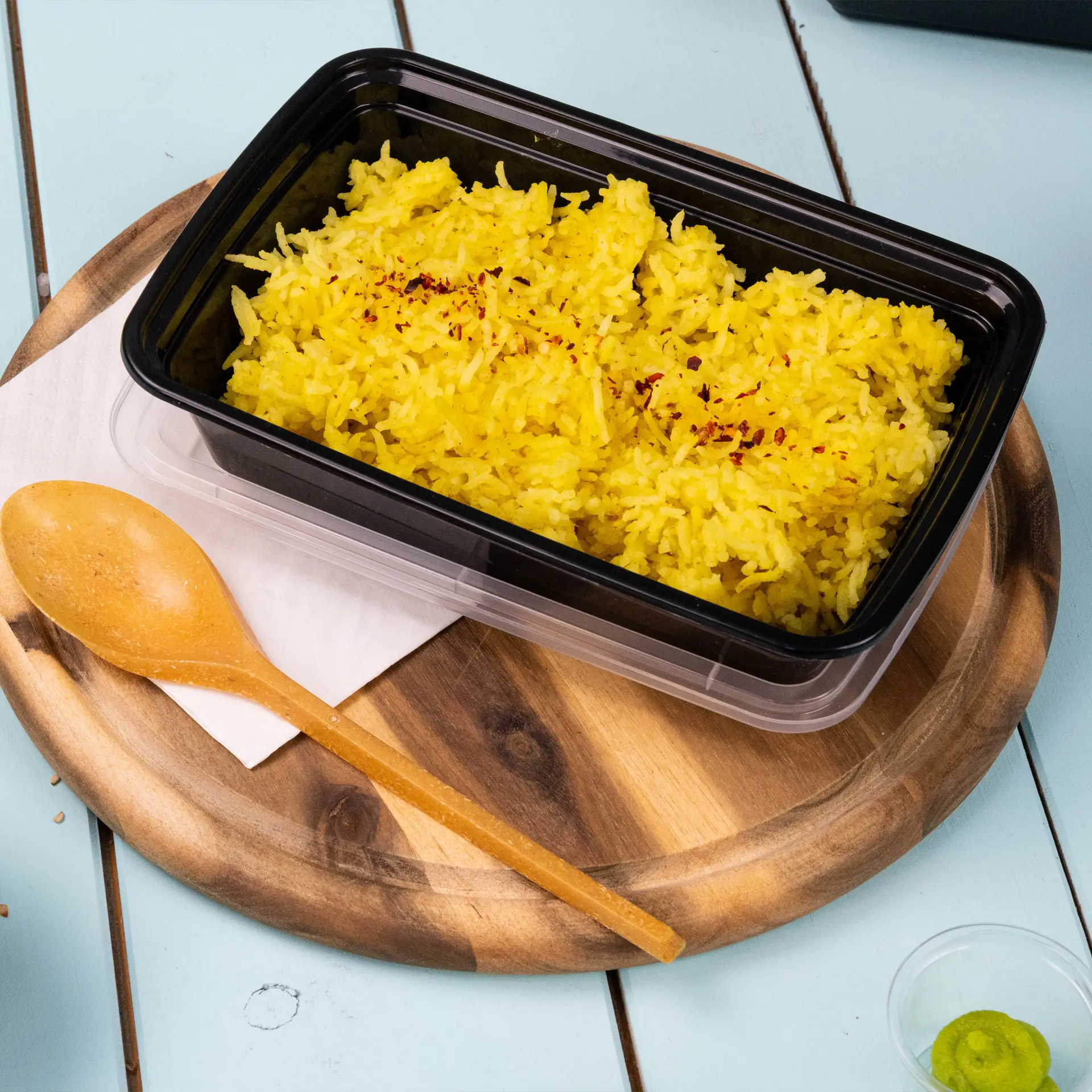 One black rectangular reusable food container filled with yellow rice, wooden spoon beside it on a round wooden platter.