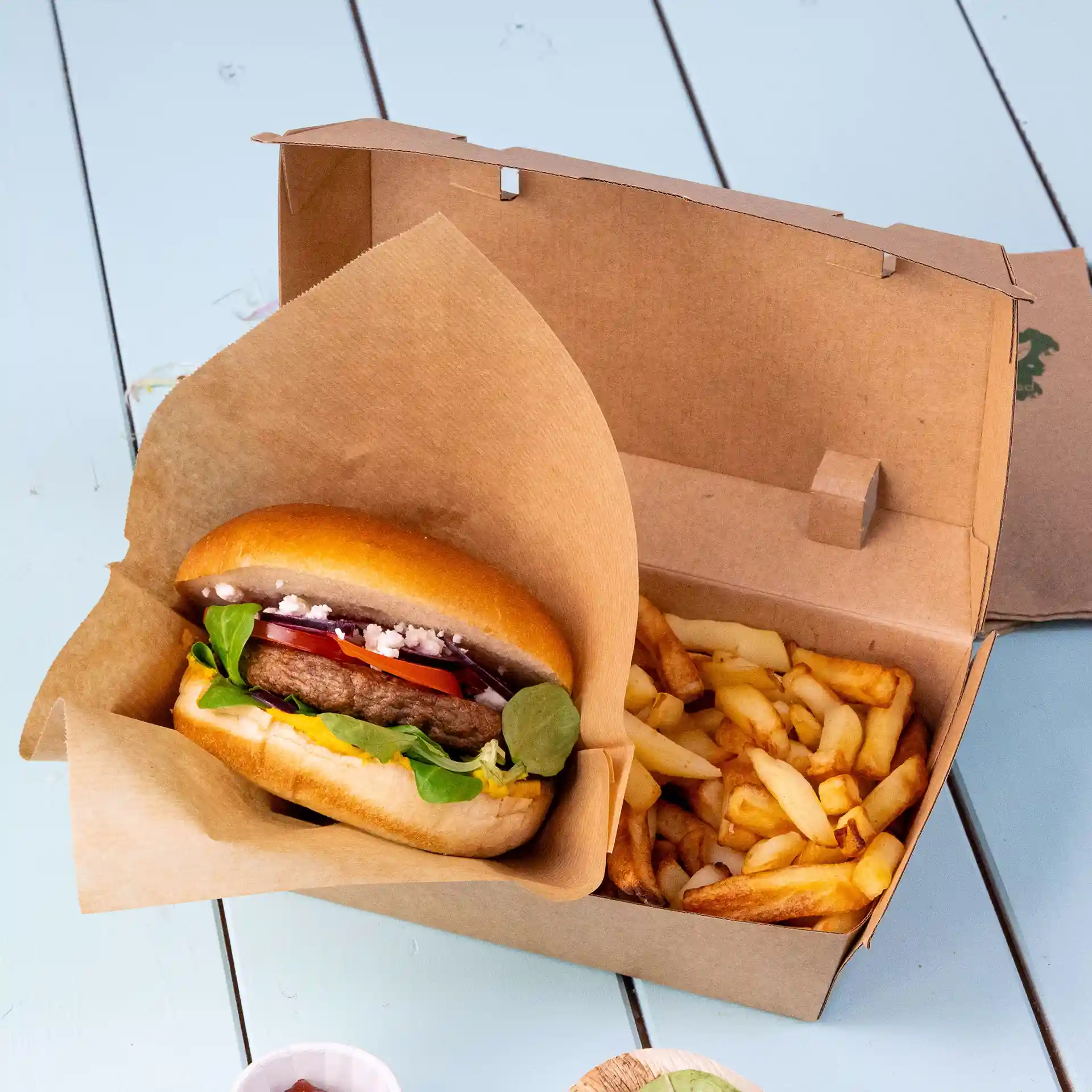 Braune Pappschachtel mit einem Hamburger und Beilagen, auf einer hellblauen Oberfläche.