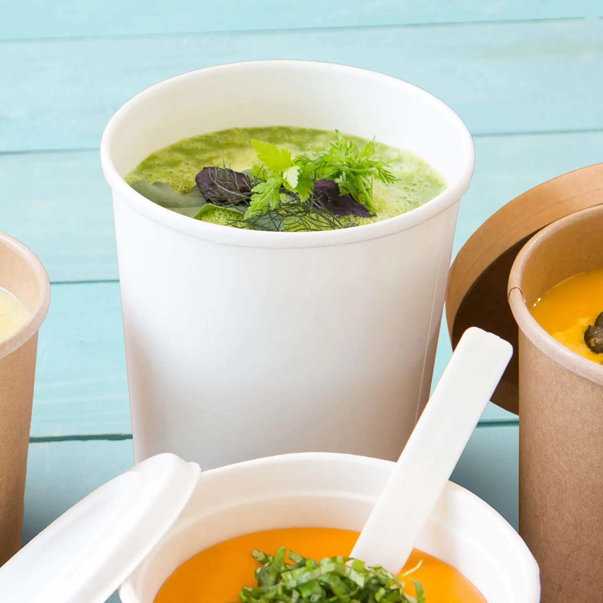 Weißer biologisch abbaubarer Suppenbecher mit grüner Suppe und Kräutern, platziert mit anderen Suppenbehältern auf einem türkisfarbenen Hintergrund.
