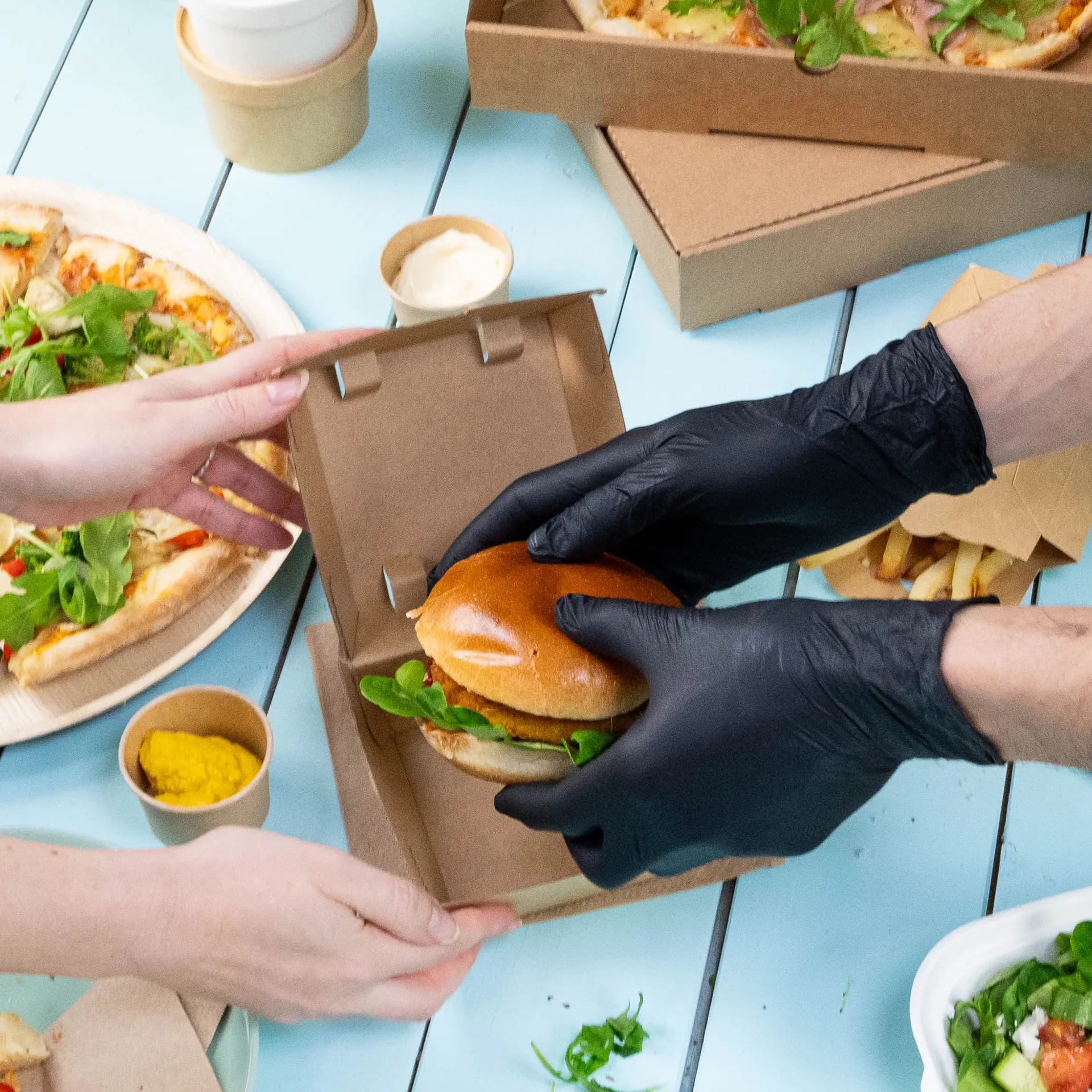 A person wearing black nitrile gloves hands a boxed burger to another person, with pizza and condiments visible nearby.