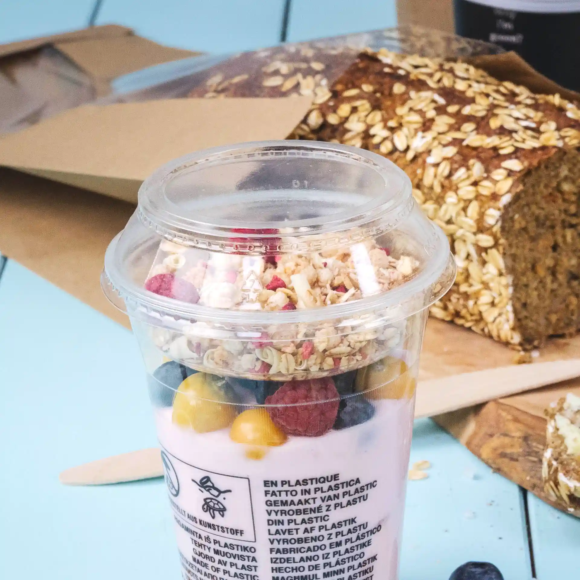 Transparenter Becher gefüllt mit Joghurt, Granola und Früchten, im Hintergrund ein Laib Haferbrot.