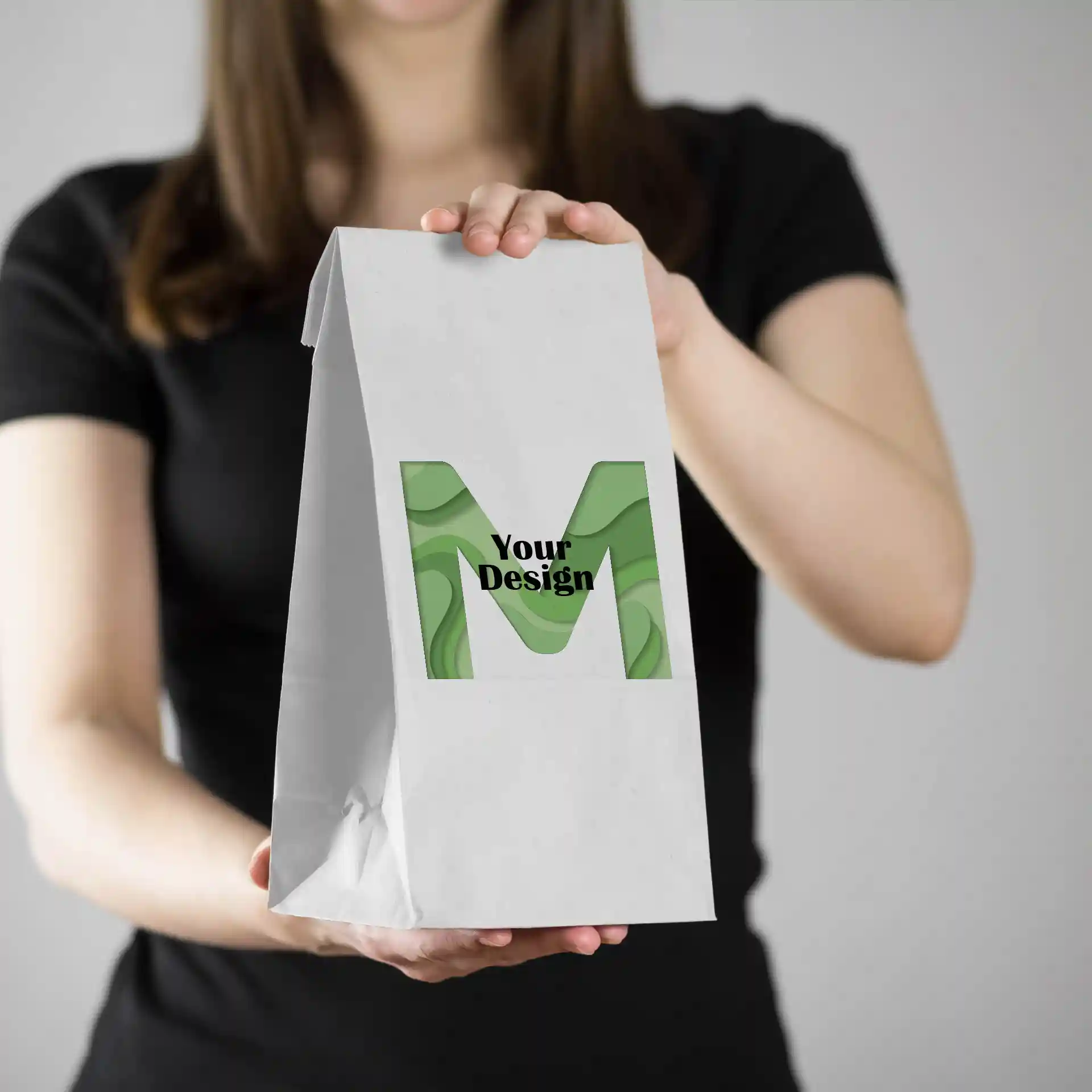 White printed paper bag held by a person, featuring a green design with the letter 'M' and the text 'Your Design'.