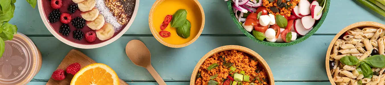 Bunte Schalen mit verschiedenen Lebensmitteln, darunter Obst, Pasta, Salat und Getreide, auf einem Holztisch angeordnet.