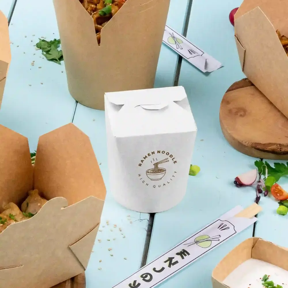 White takeout box labeled "Ramen Noodle High Quality" beside brown takeout boxes on a blue surface.