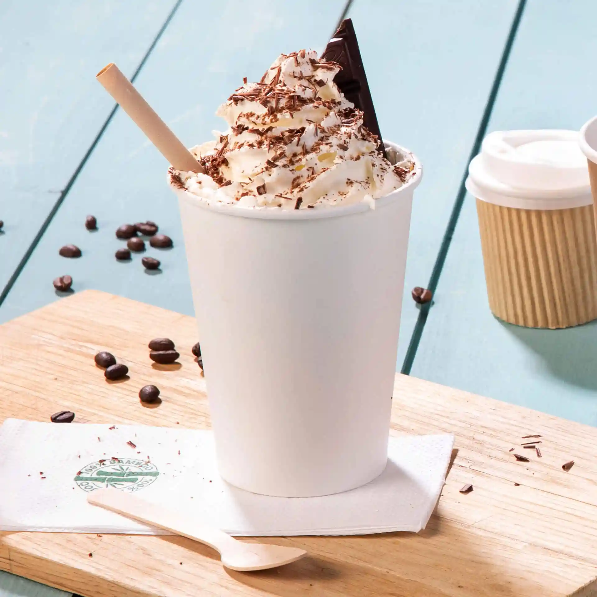 Weißer Becher mit Sahne, Schokoladenraspeln und einem Schokoladenstückchen oben, auf einer Holzoberfläche.
