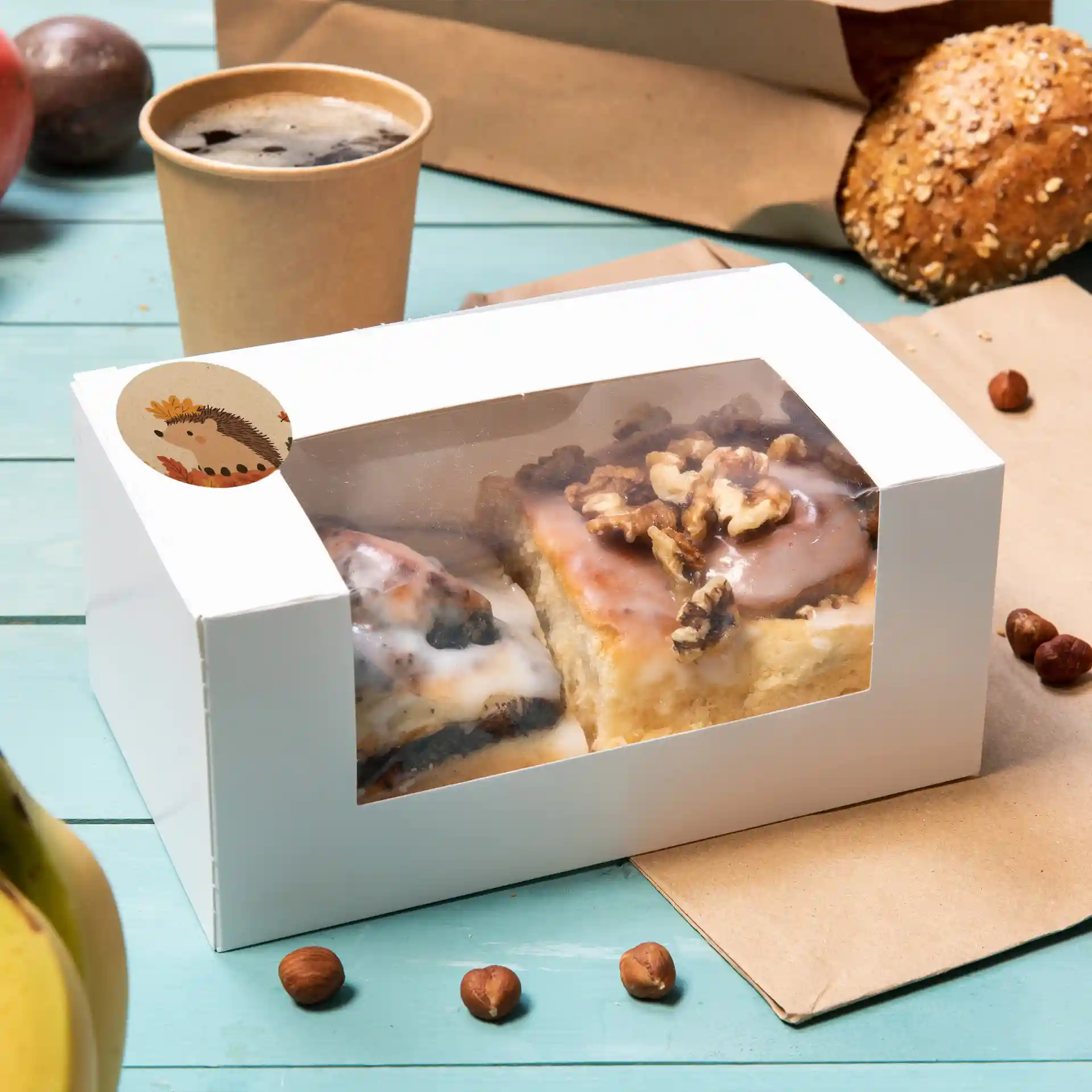White box with a clear window containing two pastries topped with icing and walnuts, surrounded by a coffee cup and hazelnuts.