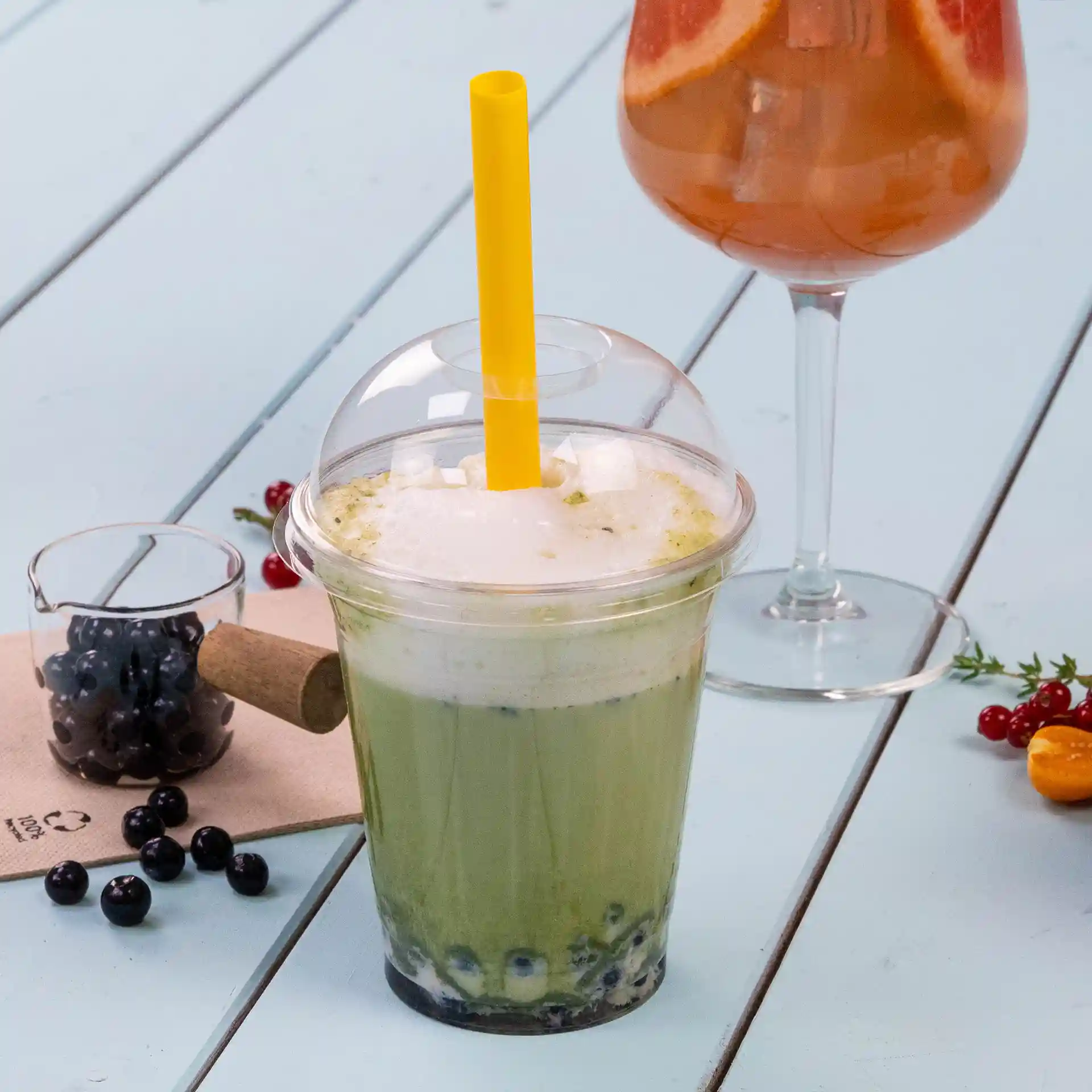 Clear cup of bubble tea with a yellow straw, featuring green tea and tapioca pearls, alongside a glass of cocktail and blueberries.