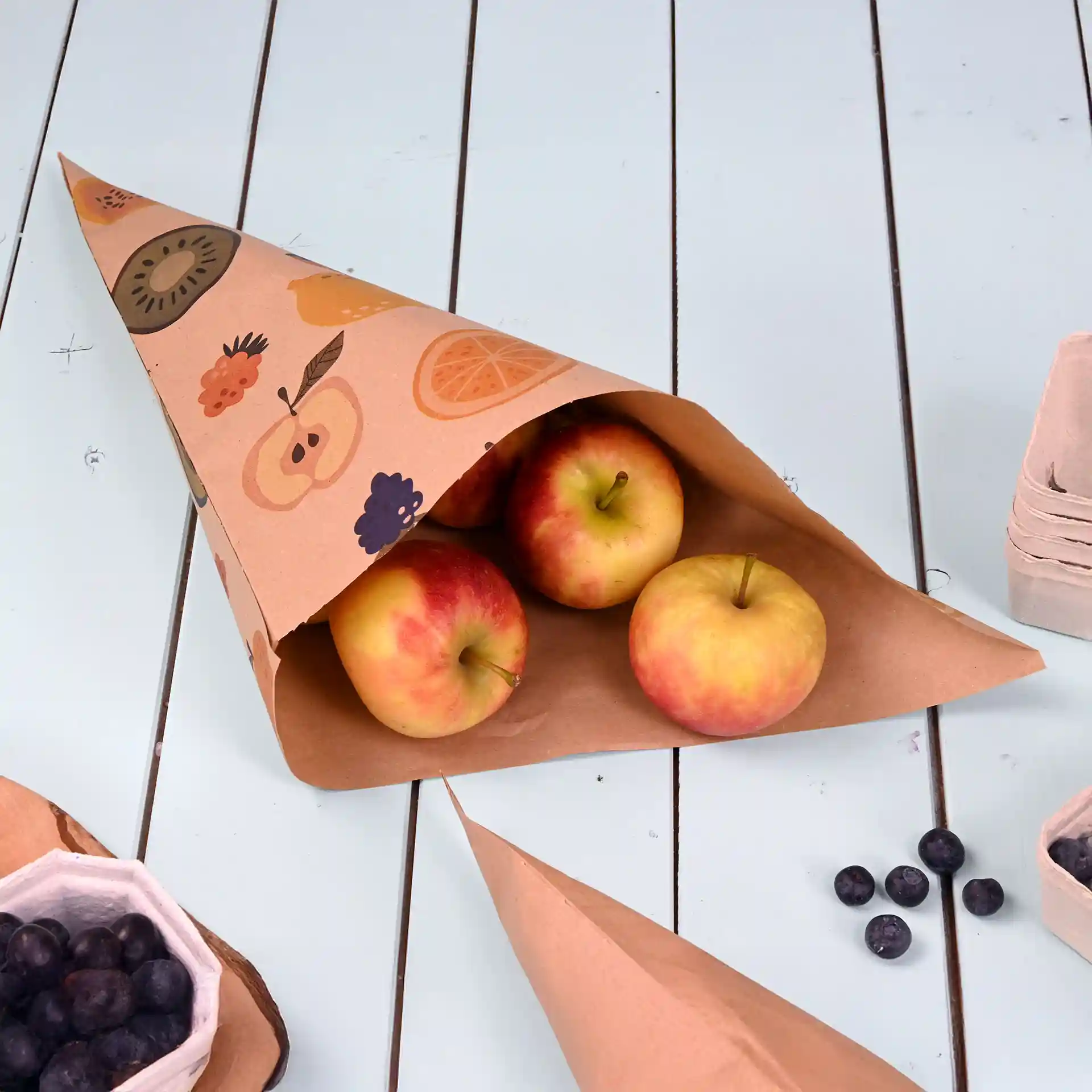 Brown paper cone bag with fruit design, containing three apples, on a light blue wooden surface.
