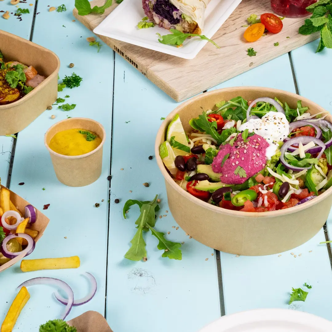 Bowl of salad with mixed vegetables, lime, and sauces, surrounded by smaller containers of sauce, fries, and herbs on a blue surface.