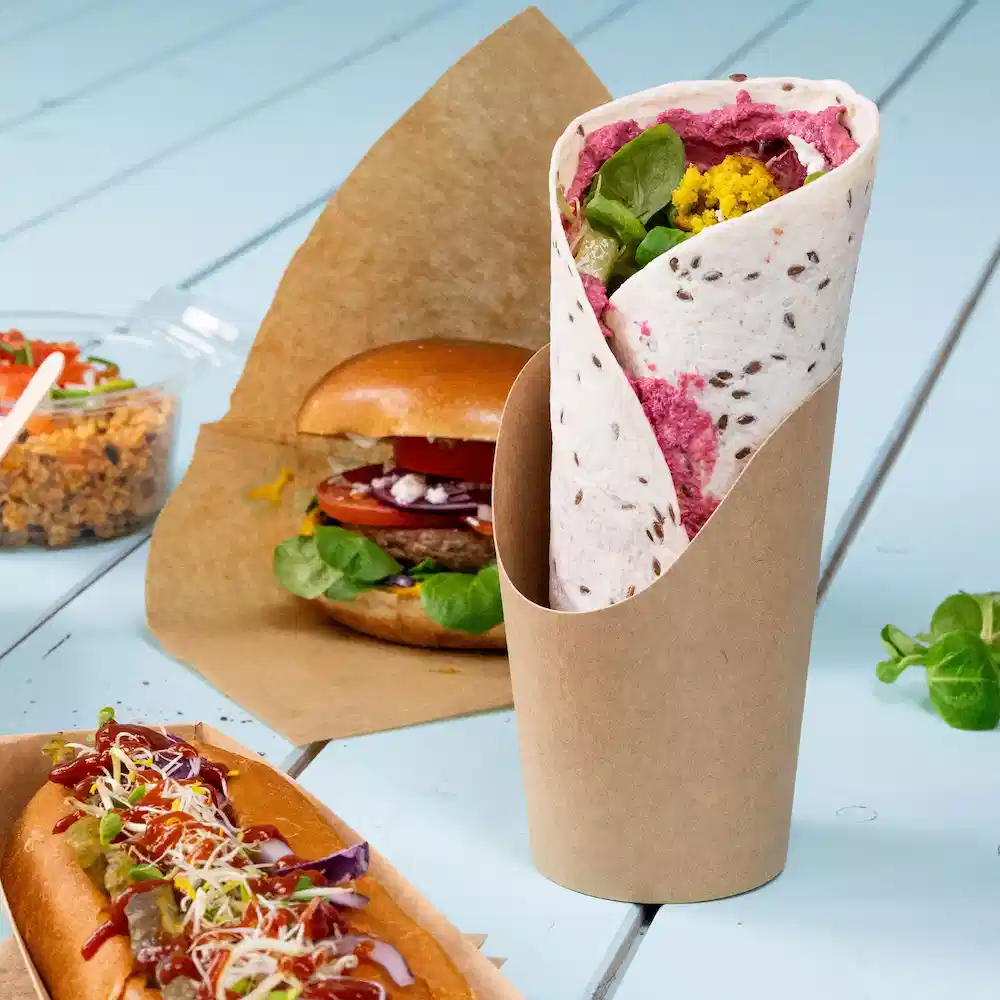 Wrap gefüllt mit Greens und Aufstrich, serviert in einem braunen Papiersack, im Hintergrund ein Burger und ein Hotdog.