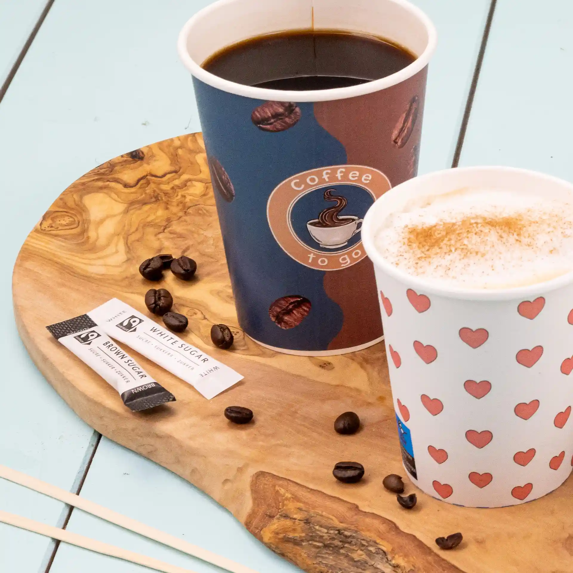 Zwei Becher auf einem Holzbrett mit Zuckersticks und verstreuten Kaffeebohnen.