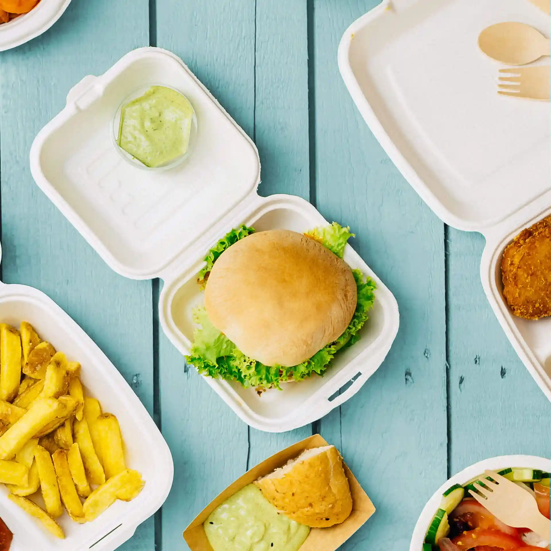 Bild einer Burgerbox aus Zuckerrohr mit einem Burger, Salat, einem kleinen Behälter mit Sauce und verschiedenen Lebensmitteln in der Nähe.