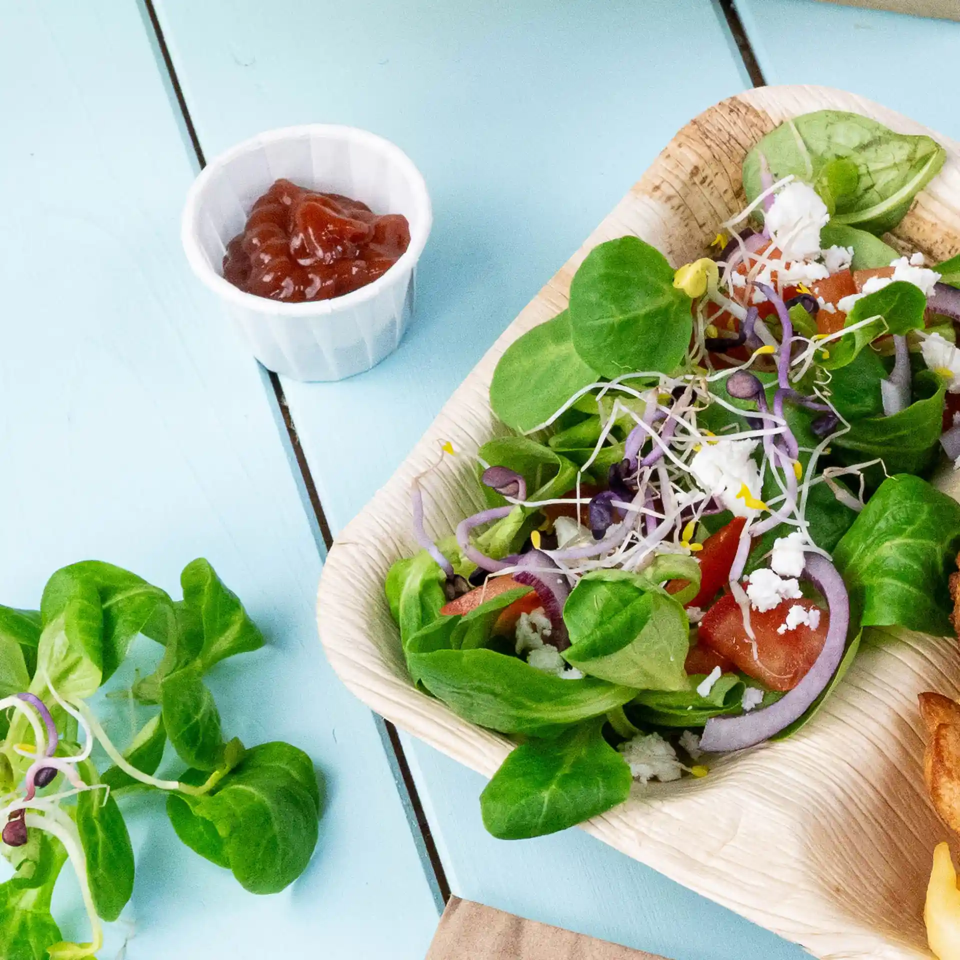 Salat mit gemischtem Grün, Tomaten, Zwiebeln und Sprossen in einer Palmblatt-Schüssel, dazu ein kleiner weißer Becher Ketchup.