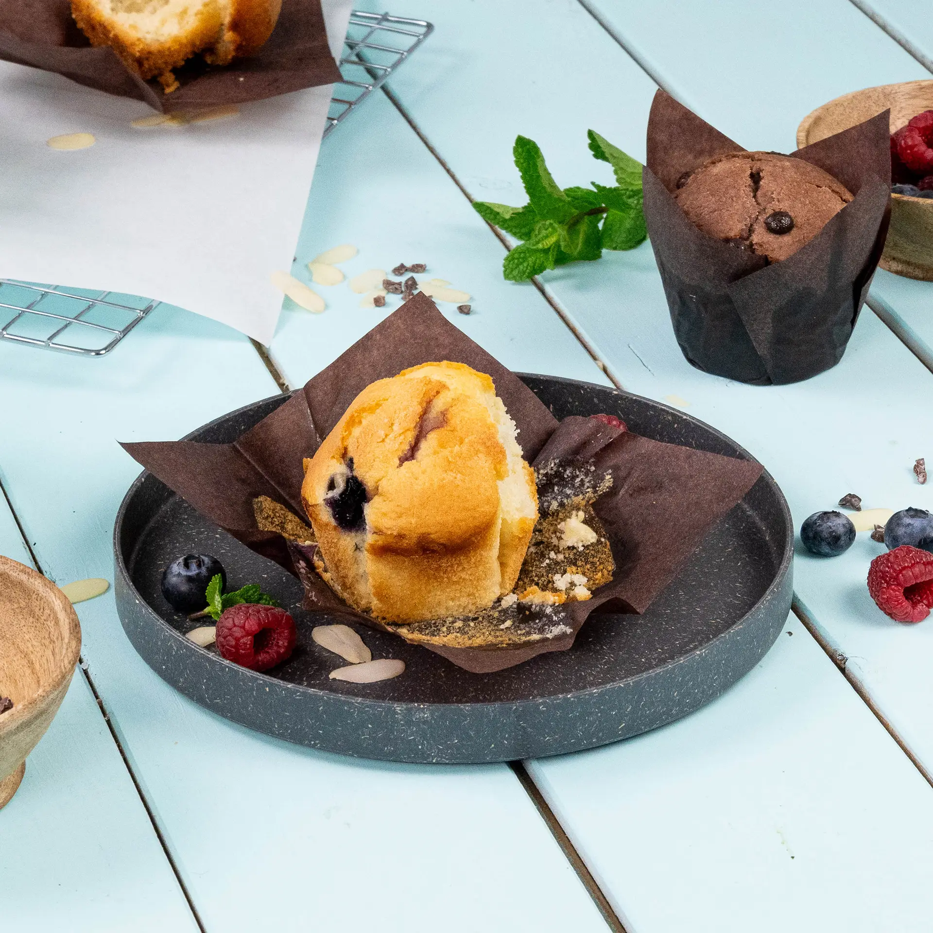 Muffin on a gray plate with brown wrapper, surrounded by raspberries, blueberries, and mint leaves.