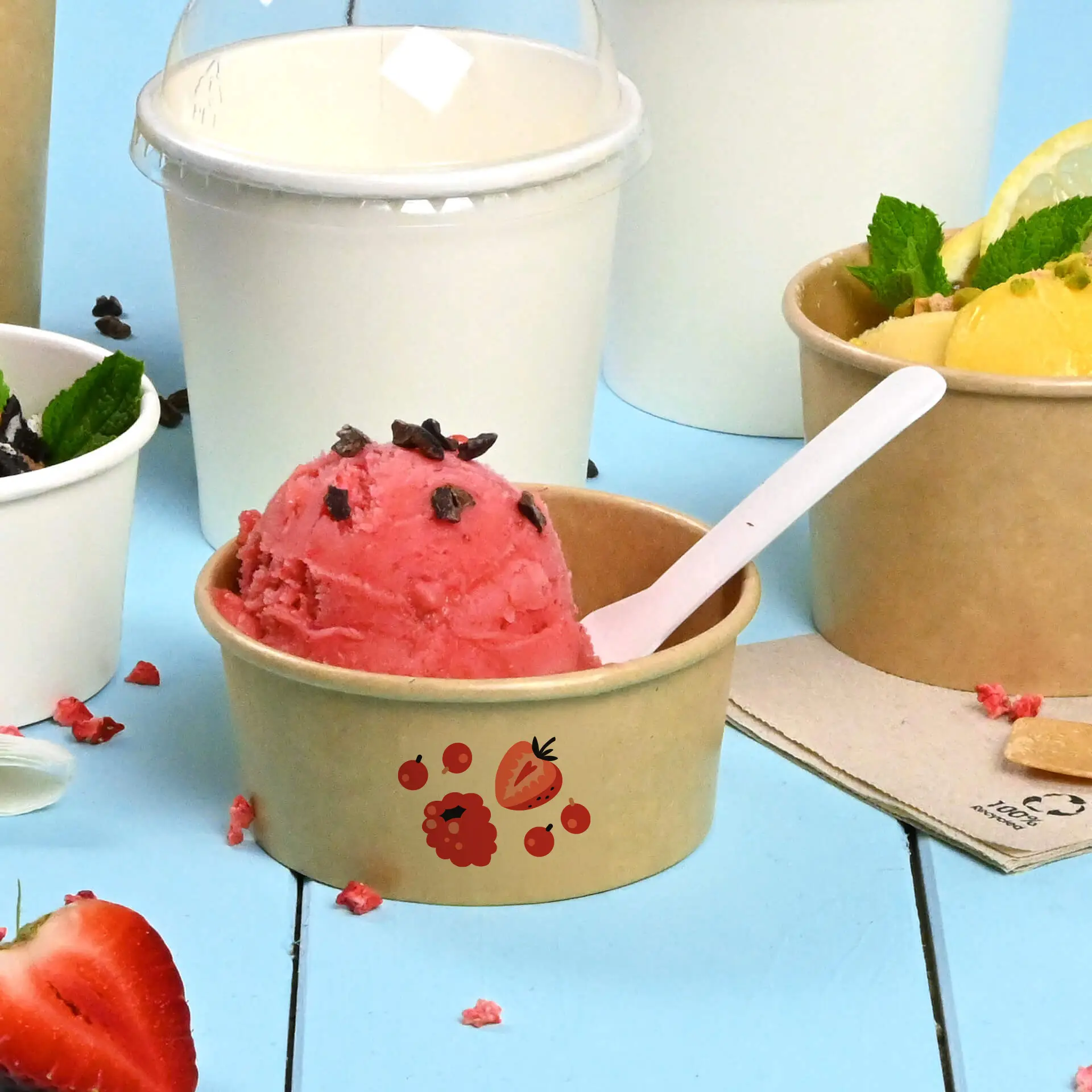 Brown ice cream cup with pink ice cream and chocolate chips, featuring printed fruit design, on a blue background.