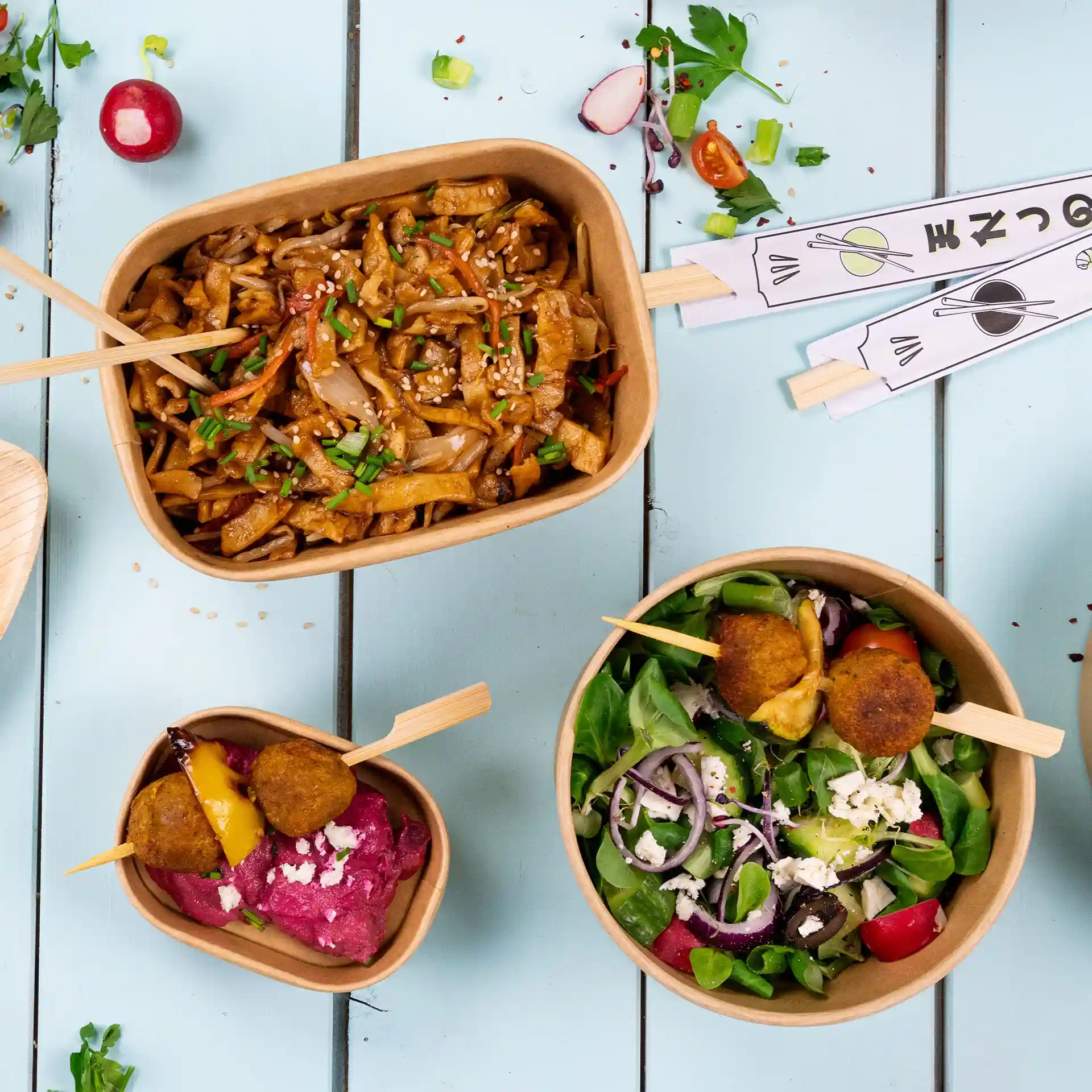 Image of three food containers: one large with noodles, a medium with salad, and a small with dip and balls, all on a blue surface.
