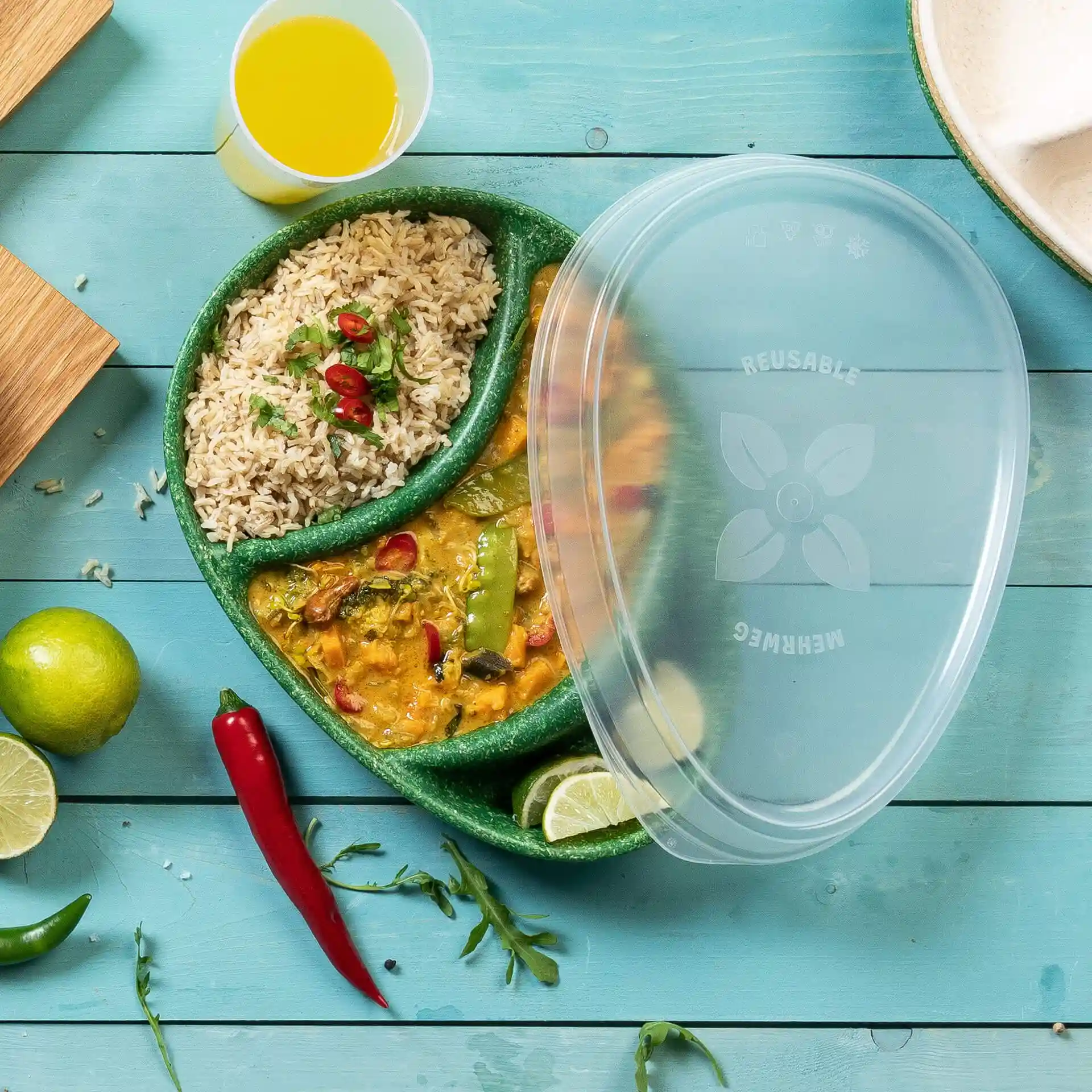 Transparent reusable plate with rice and curry, garnished with herbs and chilies, on a blue wooden surface.