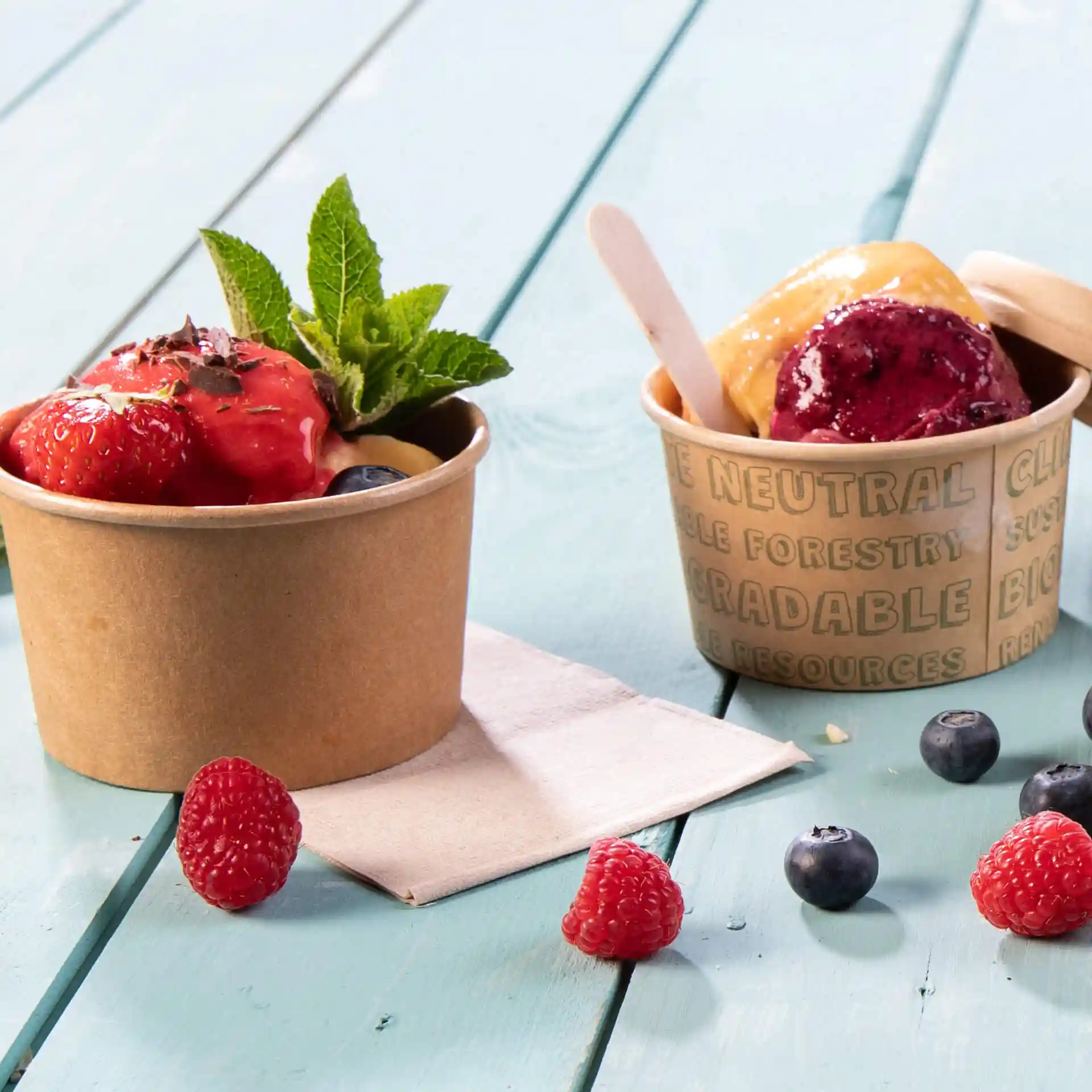 Two paper cups with ice cream, one topped with strawberries and mint, and a napkin on a blue wooden surface.