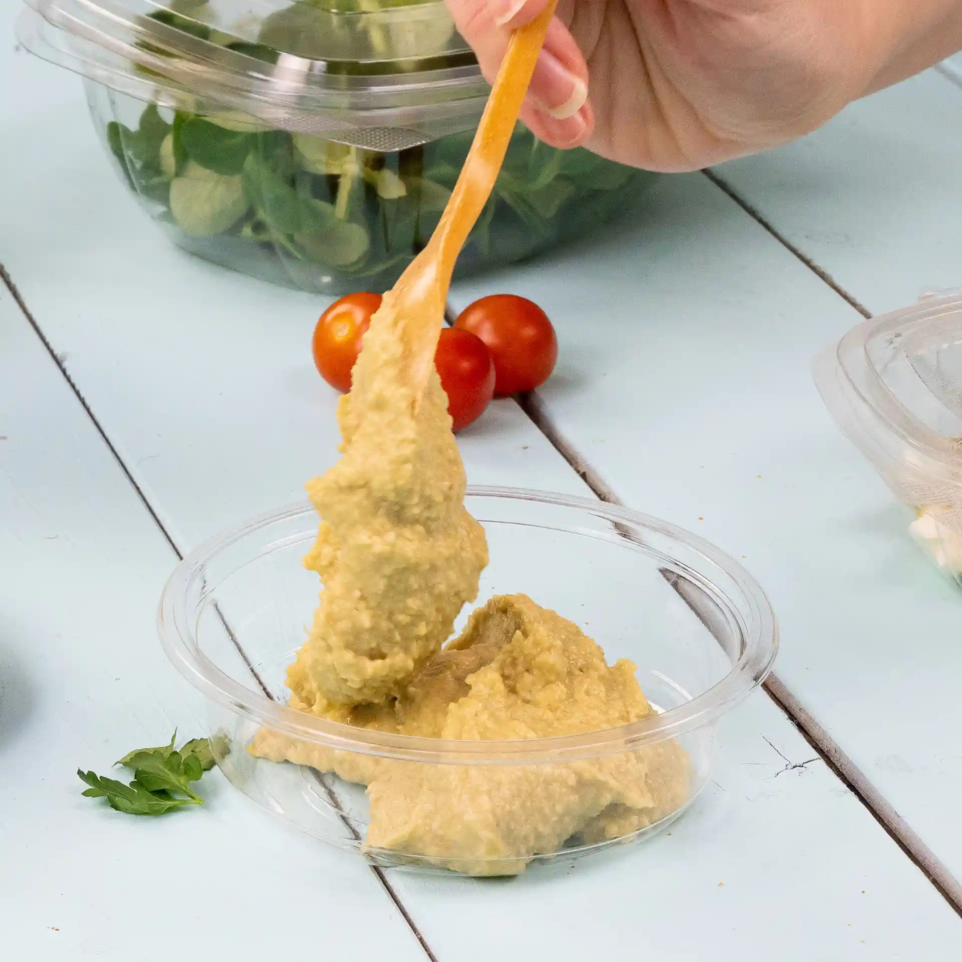A hand holding a wooden spoon scooping hummus from a clear container, with cherry tomatoes and salad in the background.