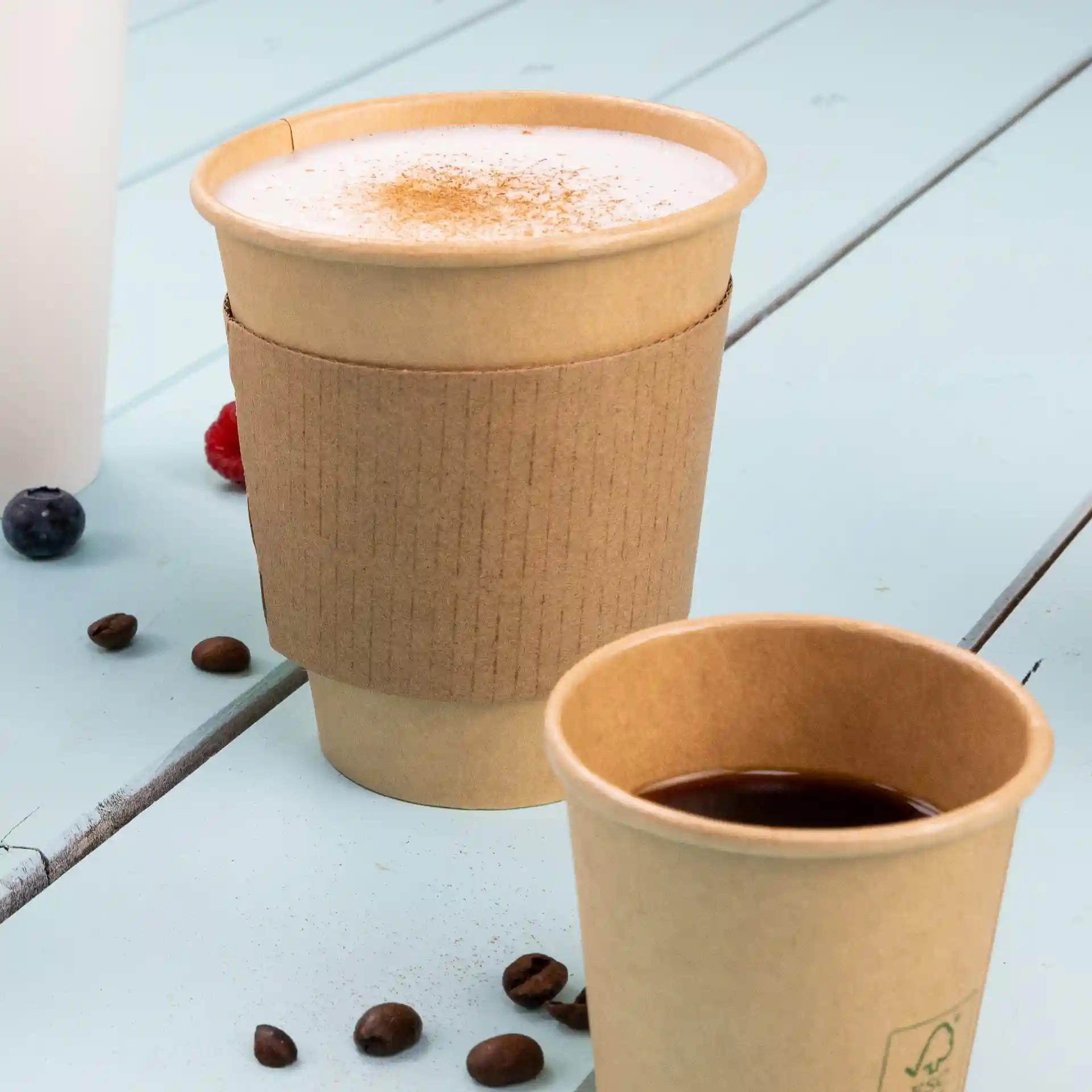 Brauner Pappbecher mit einem Kartonsleeve, gefüllt mit einem schaumigen Getränk, neben einer kleineren Tasse Kaffee und Kaffeebohnen.