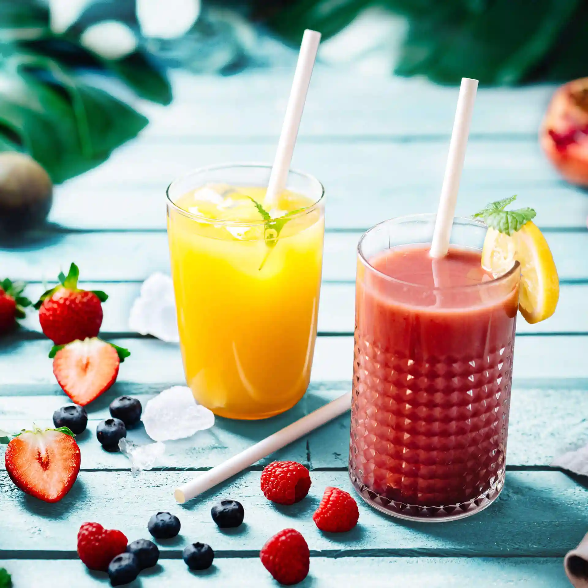 Zwei Gläser mit Getränken und weißen Papierstrohhalmen, umgeben von frischen Beeren auf einer blauen Holzoberfläche.