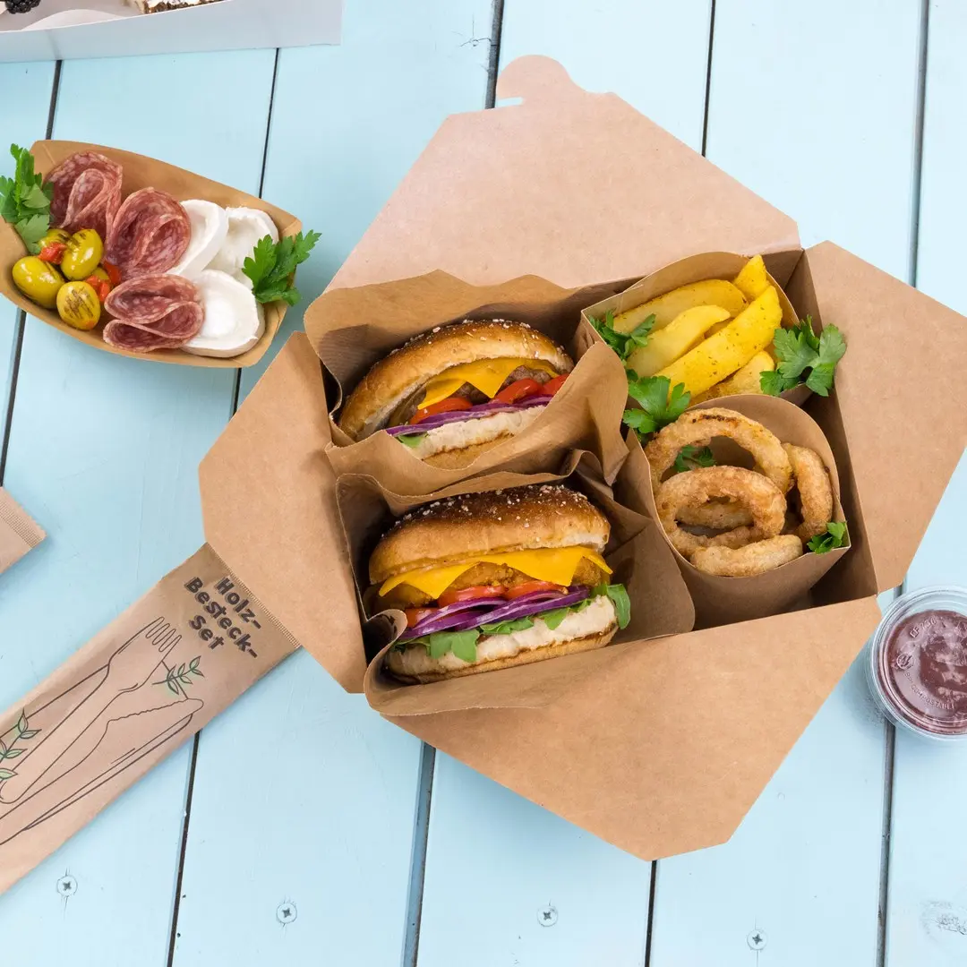 Takeaway-Box mit zwei Burgern, Zwiebelringen, Kartoffelspalten und einem kleinen Behälter mit Sauce auf einer blauen Holzoberfläche.