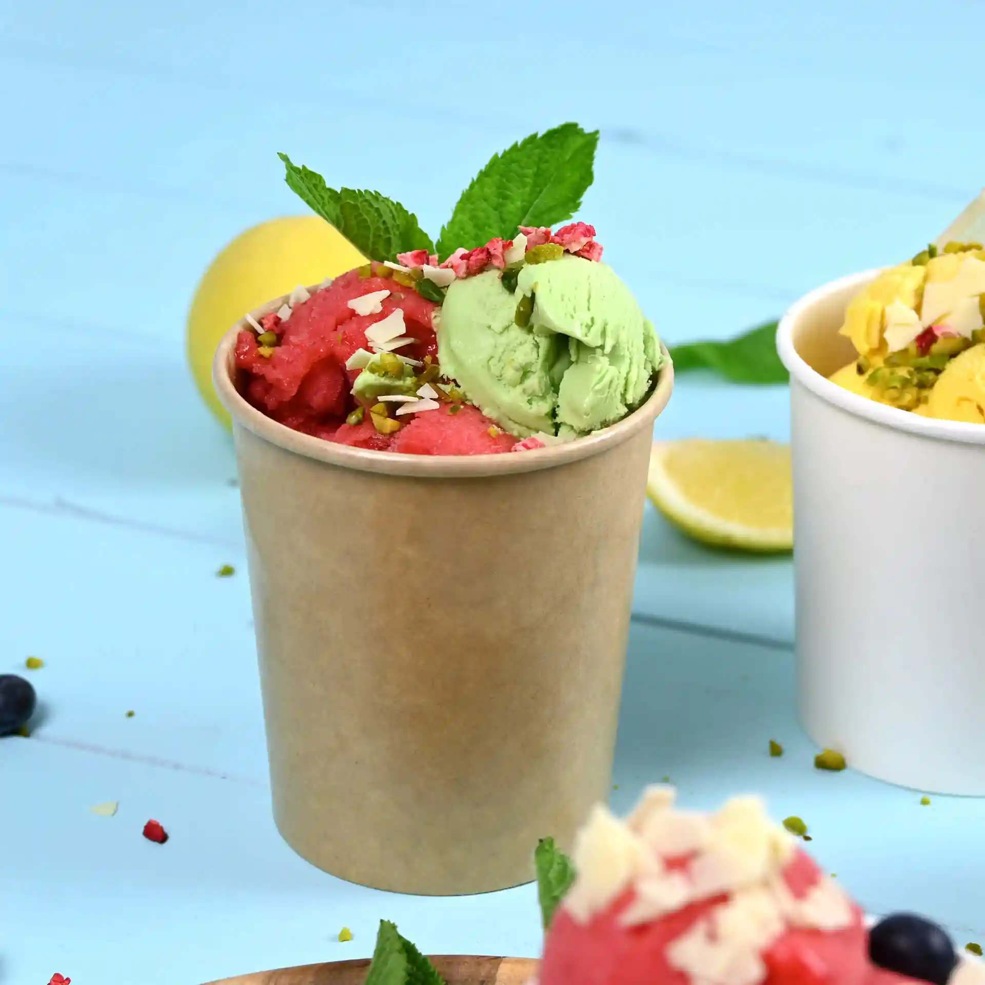 Brauner Pappbecher mit rotem und grünem Eis, garniert mit Minzblättern und Nüssen, auf blauem Hintergrund.