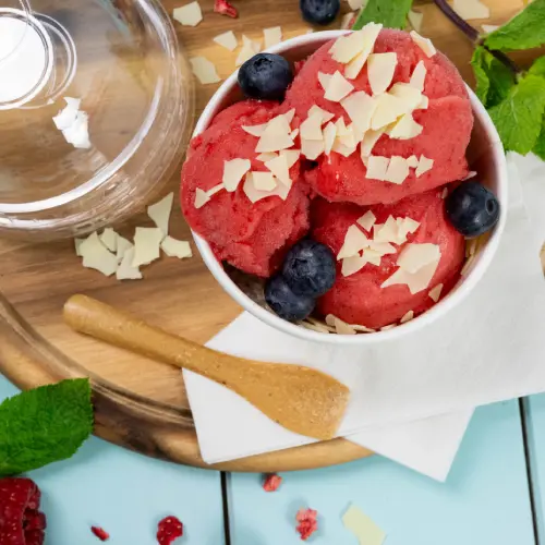 Schale mit Himbeersorbet, garniert mit Heidelbeeren und weißen Schokoladenspänen, Holzlöffel und Minzblättern auf einer Holzoberfläche.