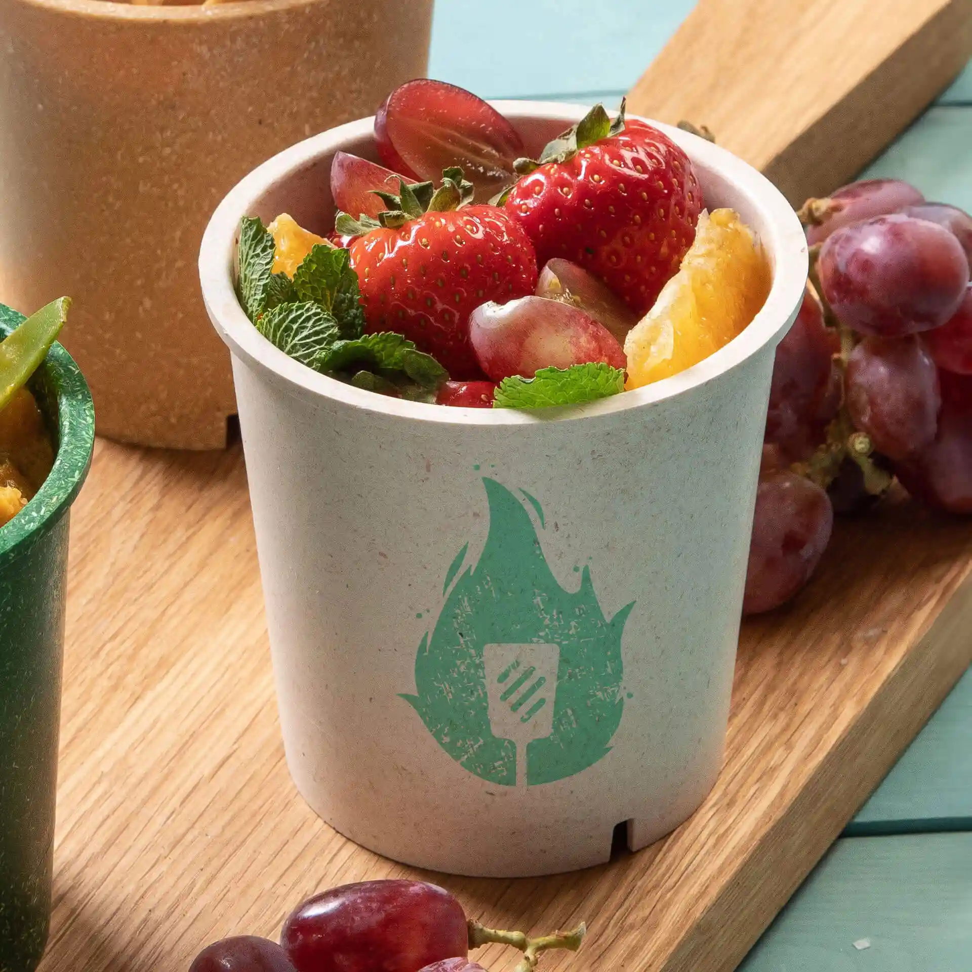 Round cream white cup filled with strawberries, grapes, and mint leaves, featuring a green flame symbol.