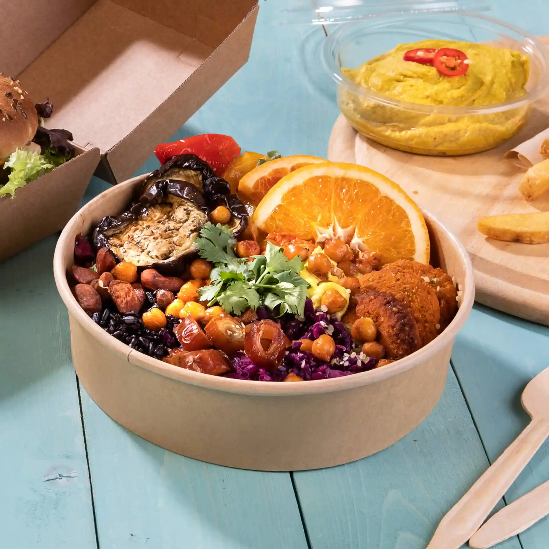 Round brown cardboard bowl filled with various vegetables, chickpeas, and orange slices, with a side of hummus and fries.
