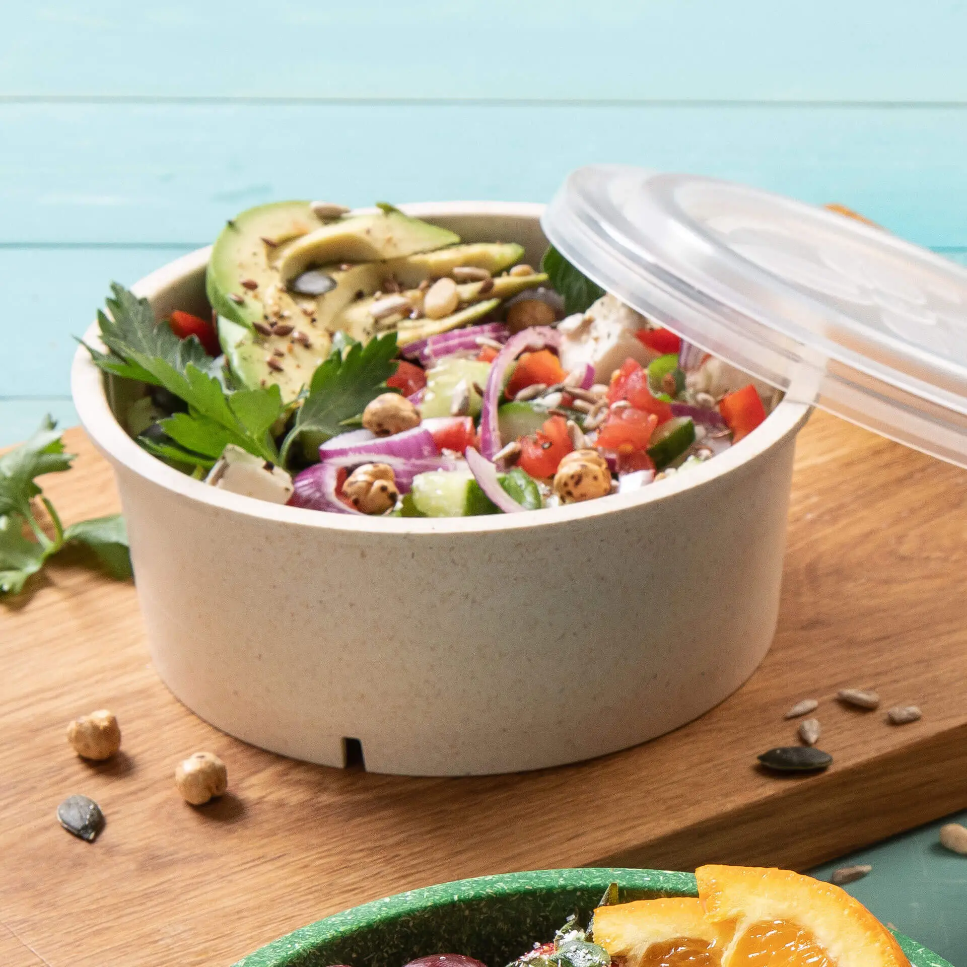 Eine Salatschüssel mit gemischtem Grün, Avocadoscheiben, Tomaten und Samen; Klarsichtdeckel oben, Holzbrett-Hintergrund.