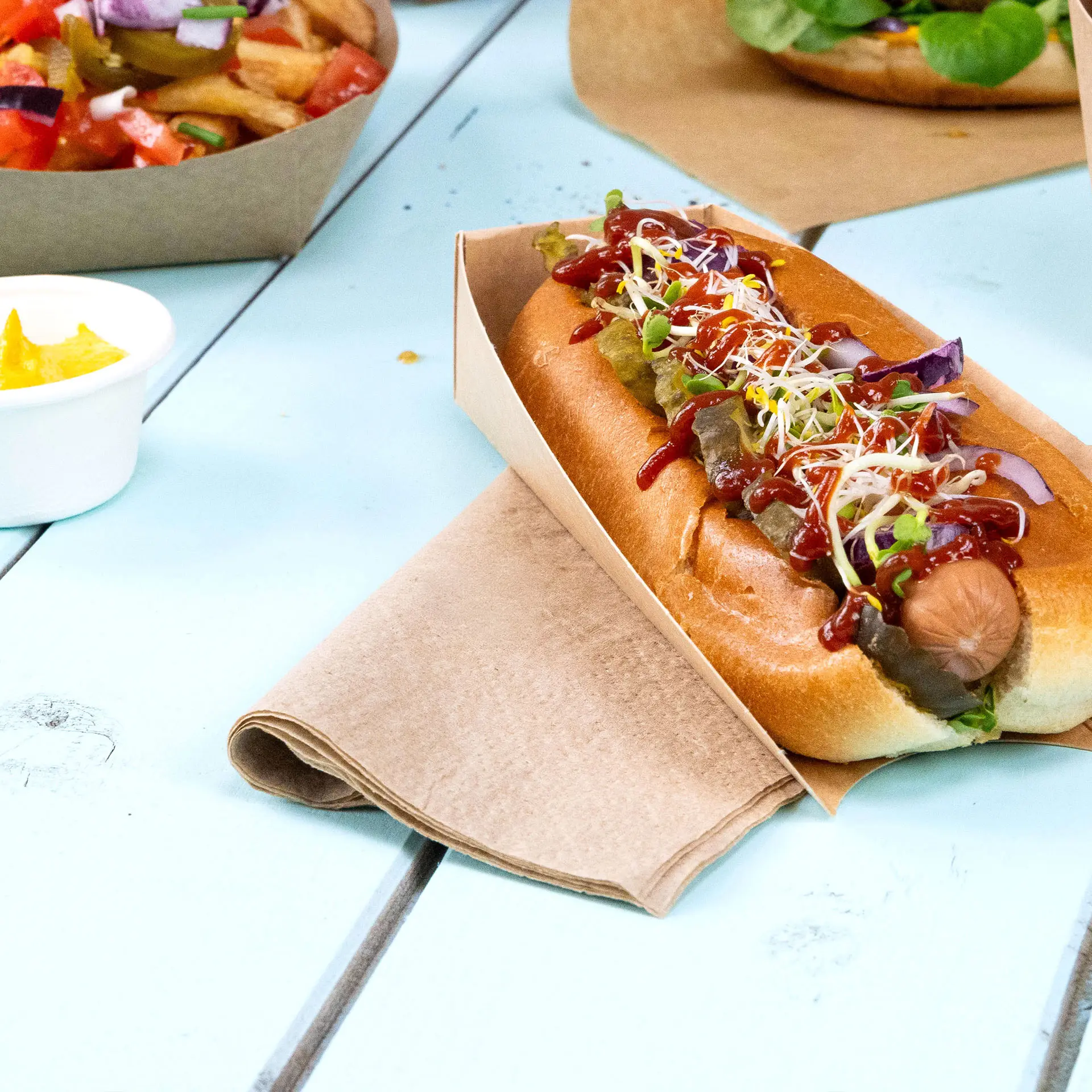 Hotdog in einem Papierhalter, garniert mit Ketchup, Senf, Gurken und Sprossen, daneben ein quadratisches ungebleichtes Serviette.