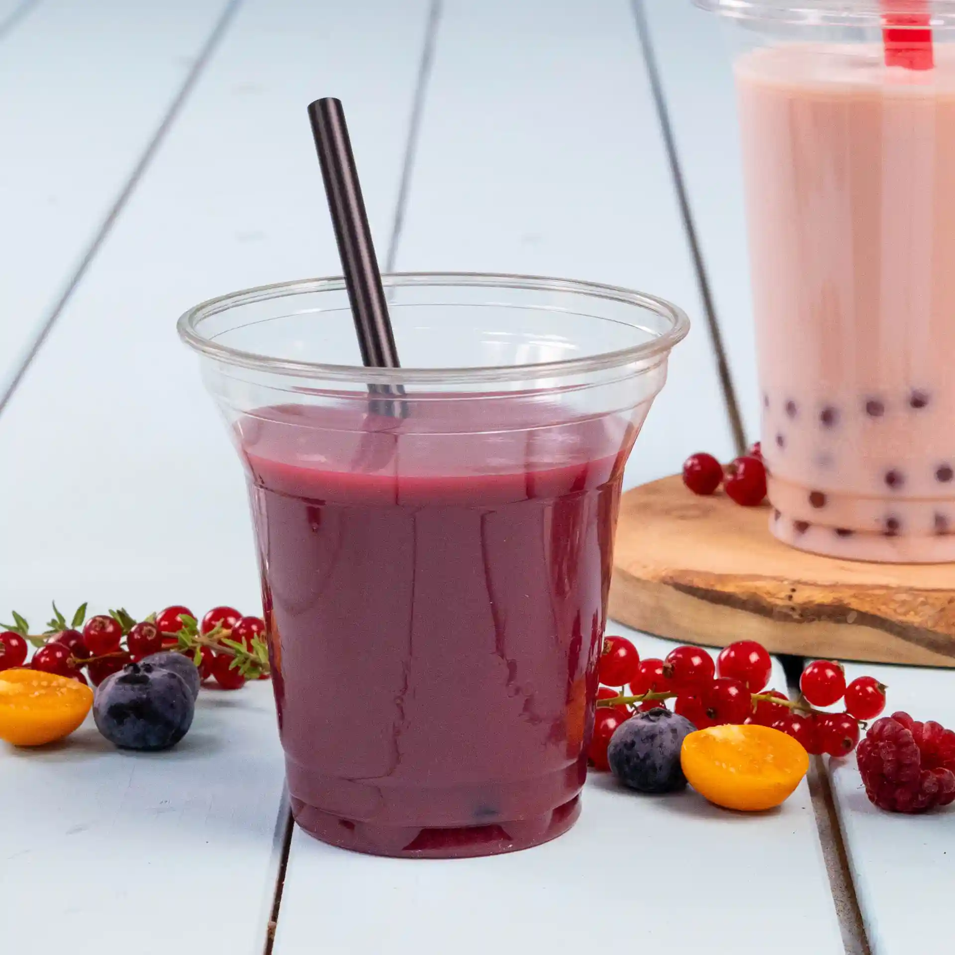Clear cup with dark purple beverage and black straw, surrounded by various berries and fruits on a light wooden surface.