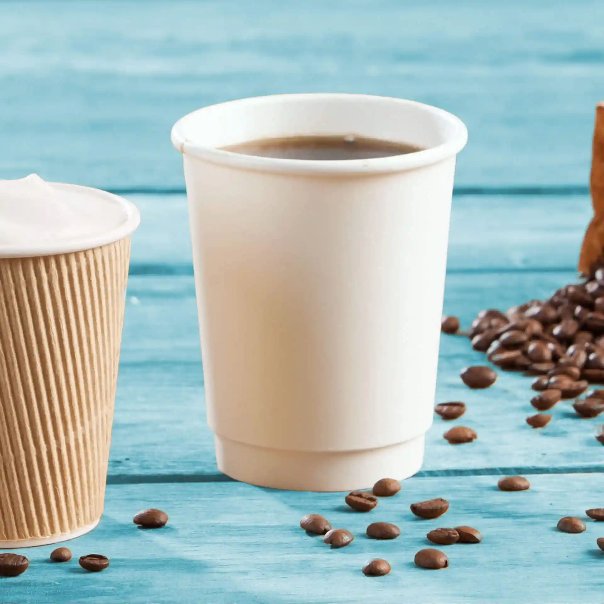 Weißer doppelwandiger Pappbecher mit Kaffee, umgeben von Kaffeebohnen und einem gerippten Papierbecher.
